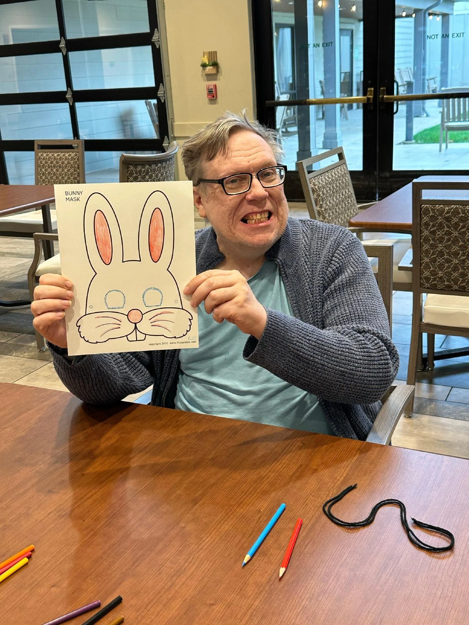 A man with glasses and gray hair smiling and showing his teeth, holding a bunny mask craft with drawn features including bunny ears, eyes, a nose, and whiskers. The setting appears to be a dining area with chairs and large windows showing an outdoor patio.