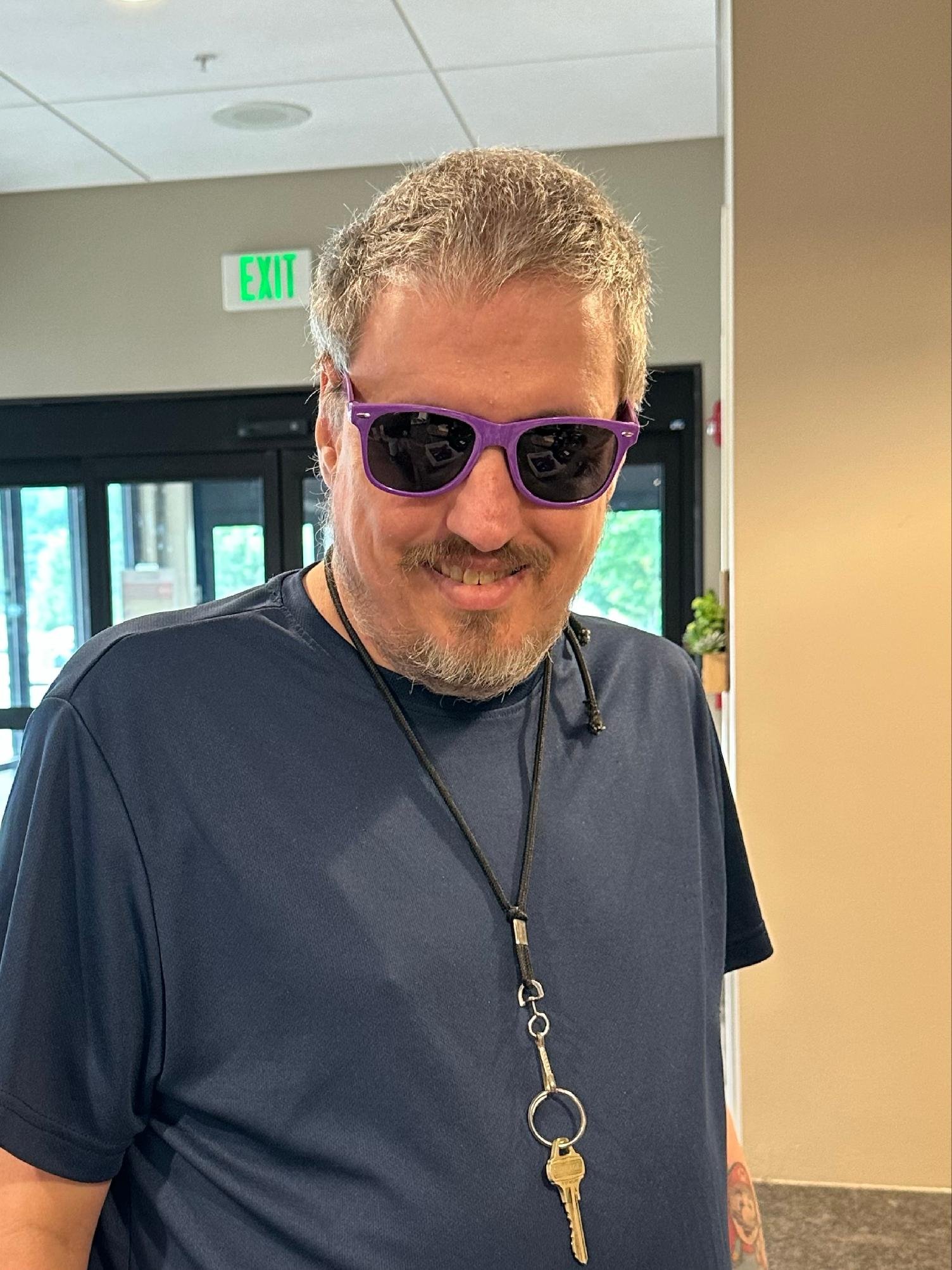 A man with short gray hair, wearing purple sunglasses, a navy blue shirt, and a necklace with a large key pendant, smiling inside a building with a green EXIT sign in the background.