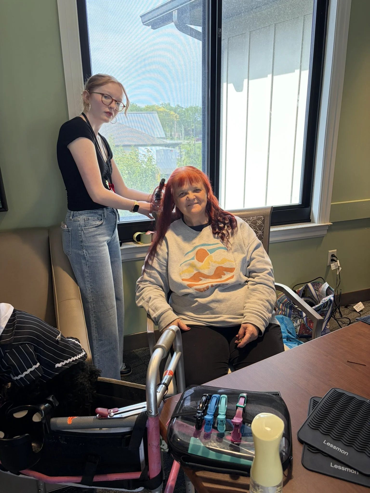 A woman with red hair sitting in a chair by a window, smiling, while another woman with glasses and light brown hair styles her hair. The table in front has various hair styling tools, and the room has green walls with sunlight coming through the win
