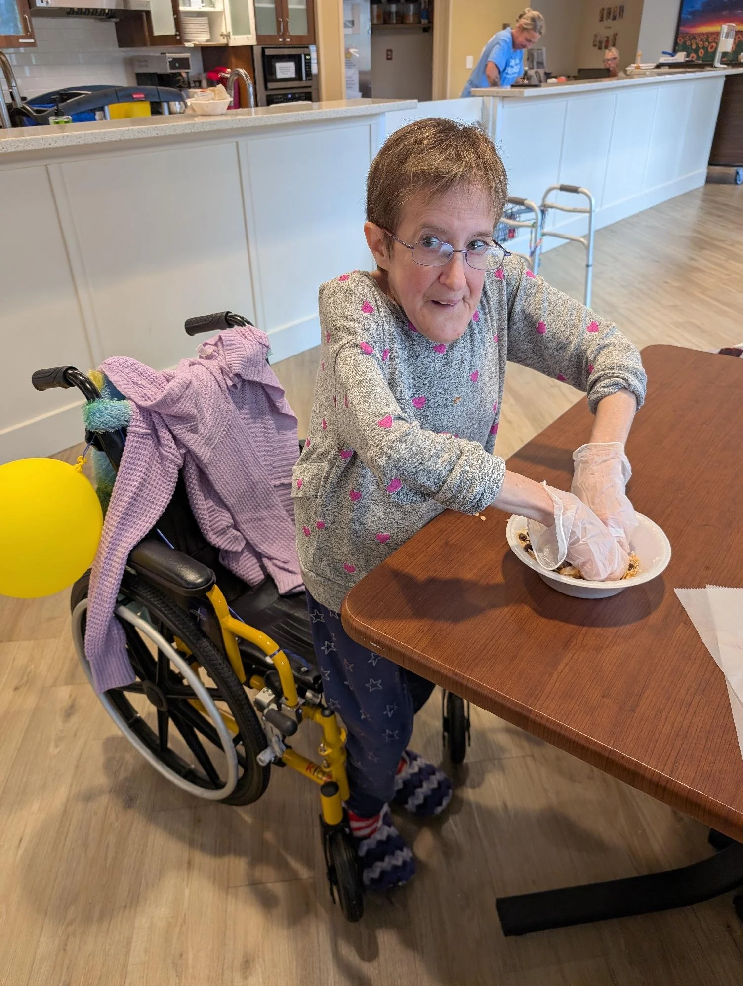 An elderly woman with short brown hair, glasses, and a grey sweater with pink hearts, sitting in a yellow wheelchair. She is wearing colorful striped socks and is eating at a wooden table with a bowl in front of her. A pink sweater or blanket is drap