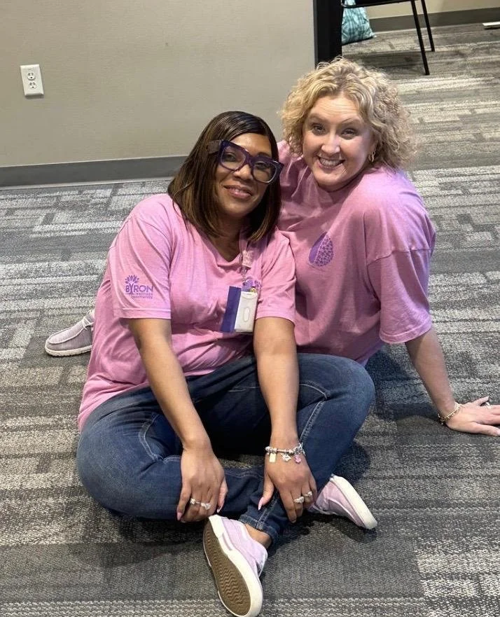 Two women sitting on the floor in pink shirts, smiling at the camera, in an indoor setting with gray carpet and plain walls.