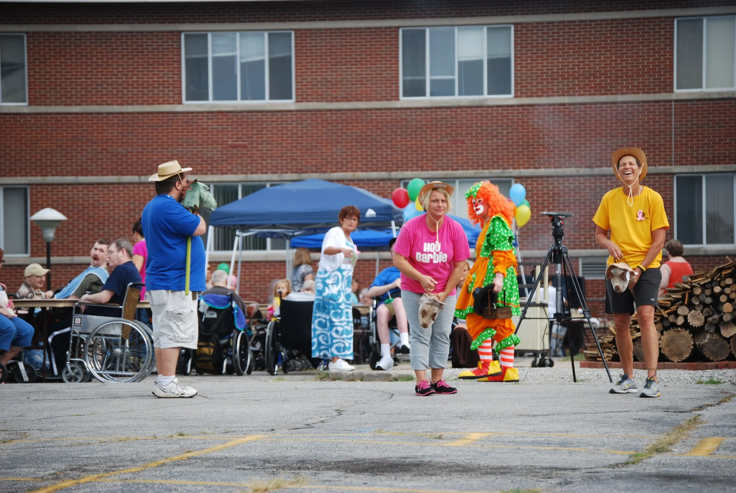 People gathered outdoors at a community event, including a woman dressed as a clown with orange curly hair and colorful costume, a woman in a pink 'HOU Barbie' shirt, and individuals in casual clothing, some in wheelchairs, with a brick building and stacked logs in the background.