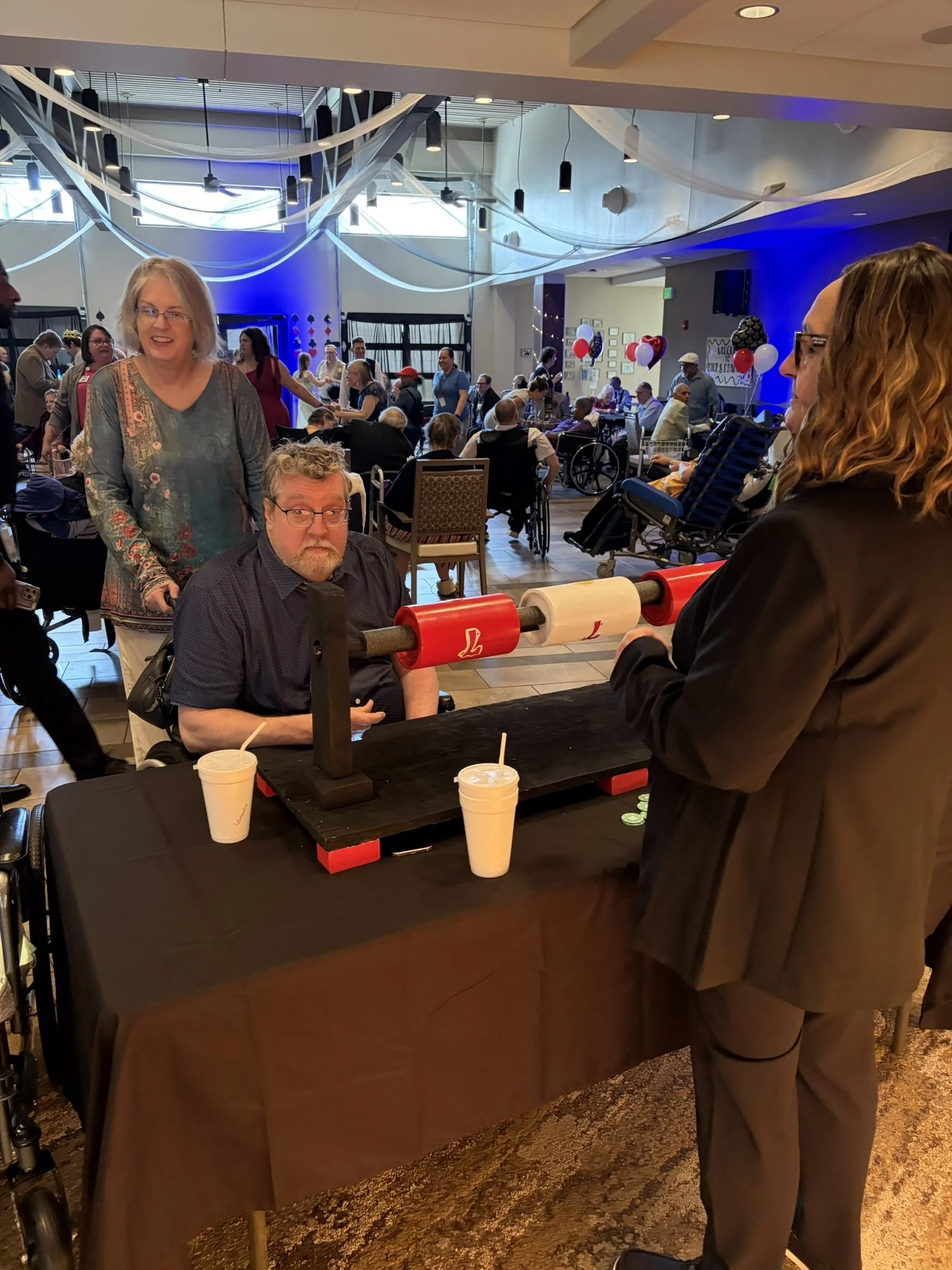 A man with glasses and a beard sitting at a table with two large cups and a paper roll on a black table. A woman standing behind him, smiling, with short gray hair and a patterned blouse. A woman with long curly hair and glasses is standing on the ri