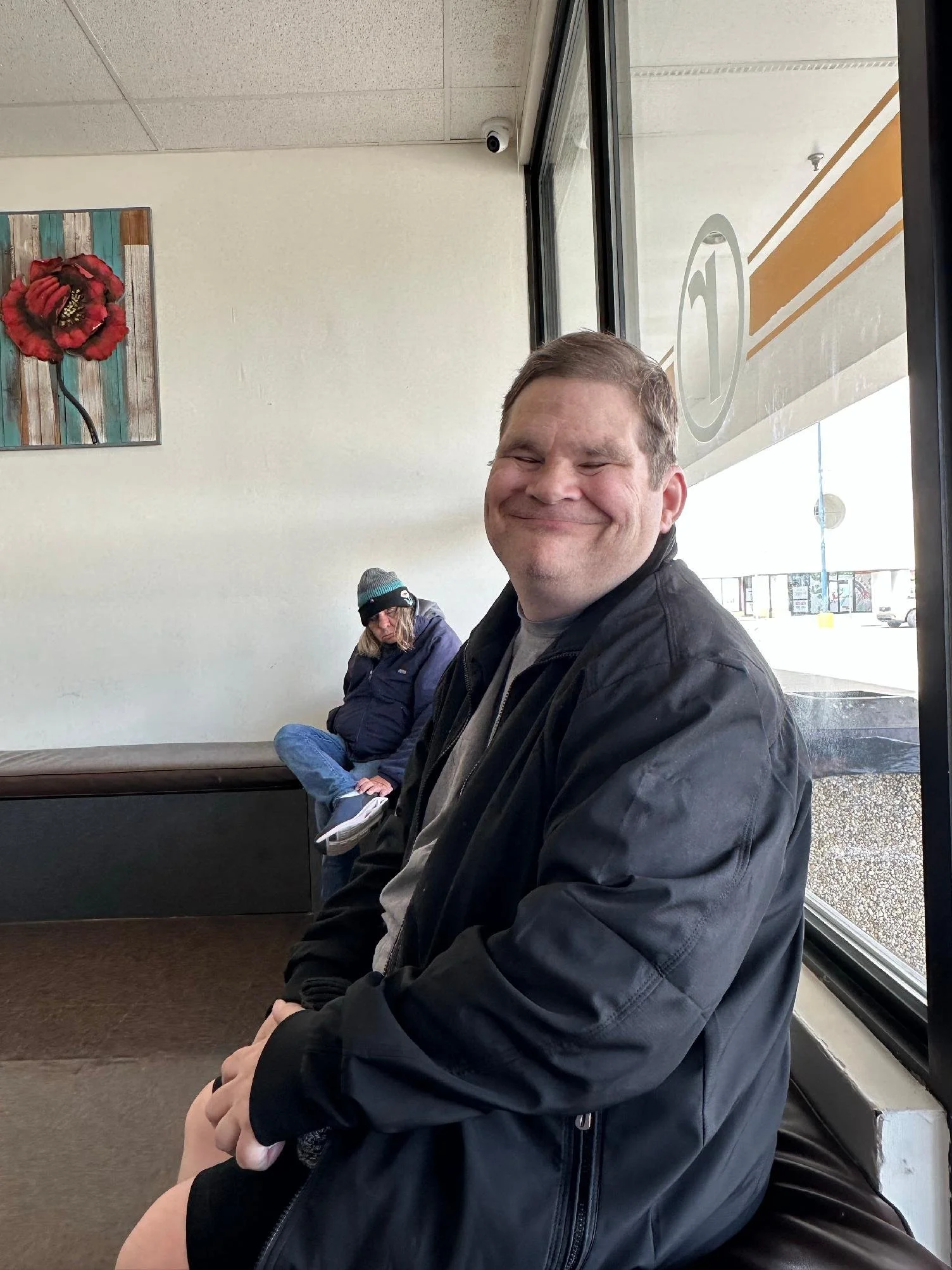 Man with short hair and fair skin smiling while sitting at a window in a waiting area, with a woman in a winter coat and hat seated behind him.
