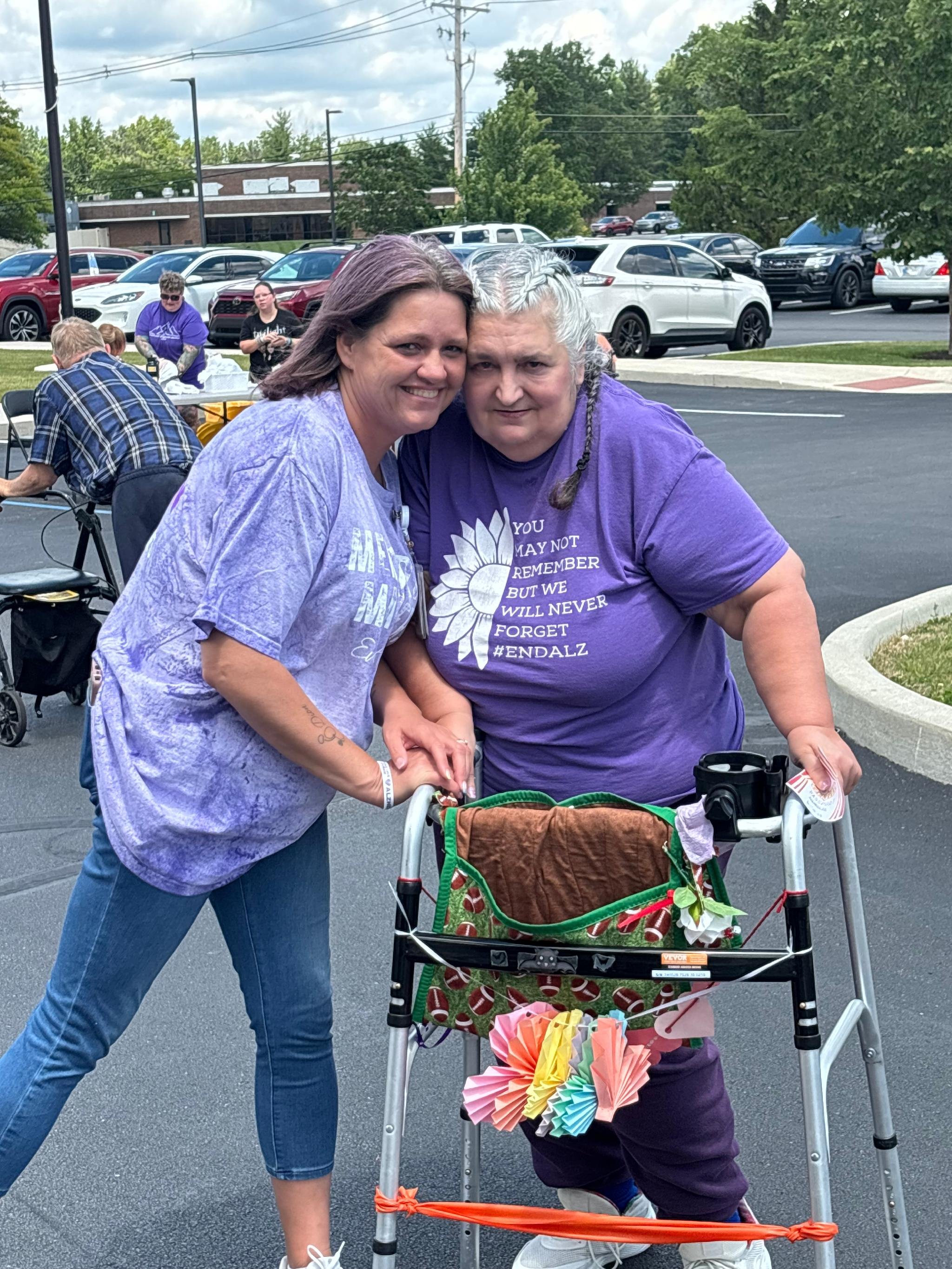 Two women standing together, one with her arm around the other, smiling at the camera. One woman is using a walker decorated with colorful paper flowers and a ribbon, outside in a parking lot with cars and trees in the background.
