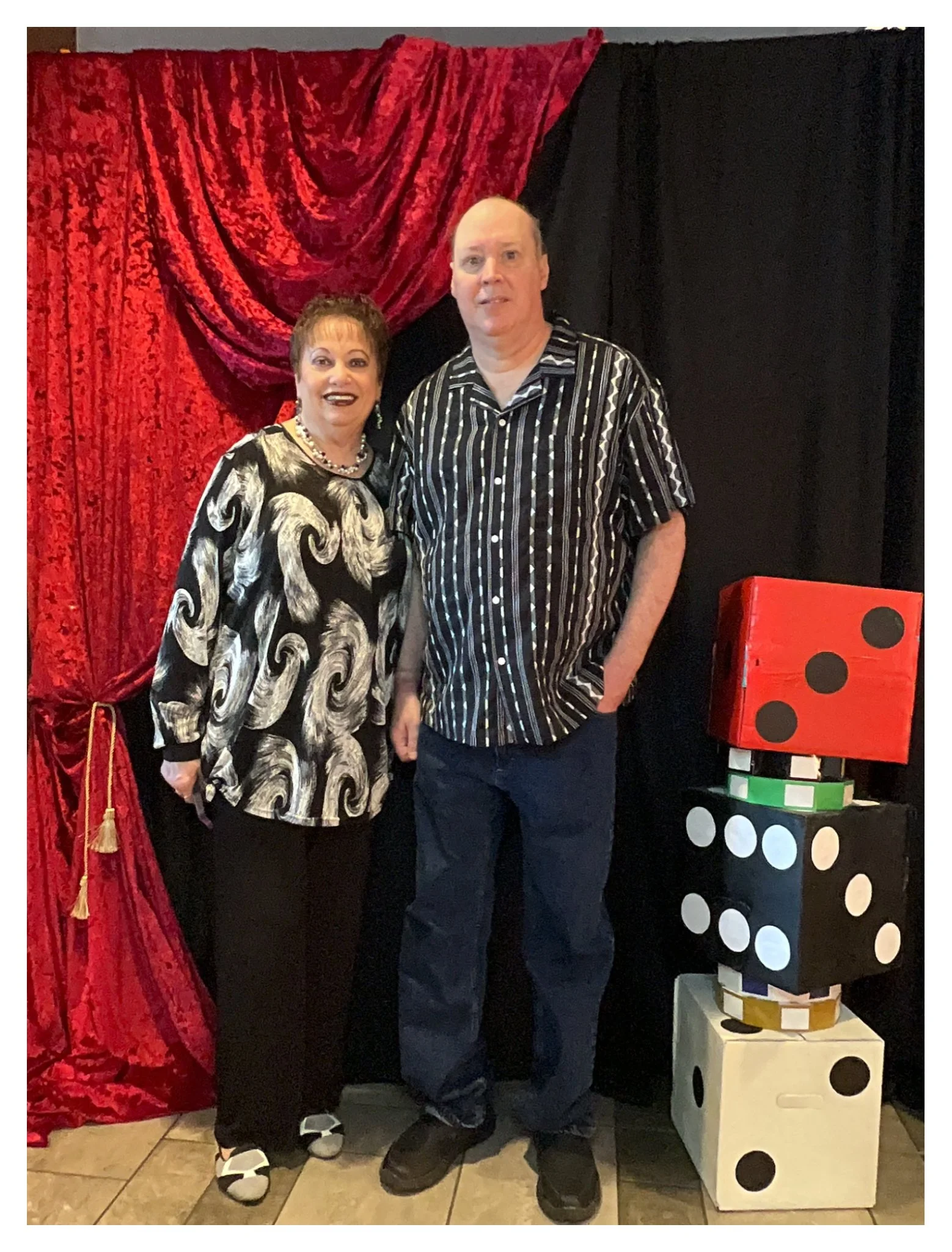 A woman and a man standing together in front of a red, textured curtain and black backdrop, with large dice props to their right. The woman is wearing a black and white swirling patterned dress, a pearl necklace, and patterned slippers. The man is dr