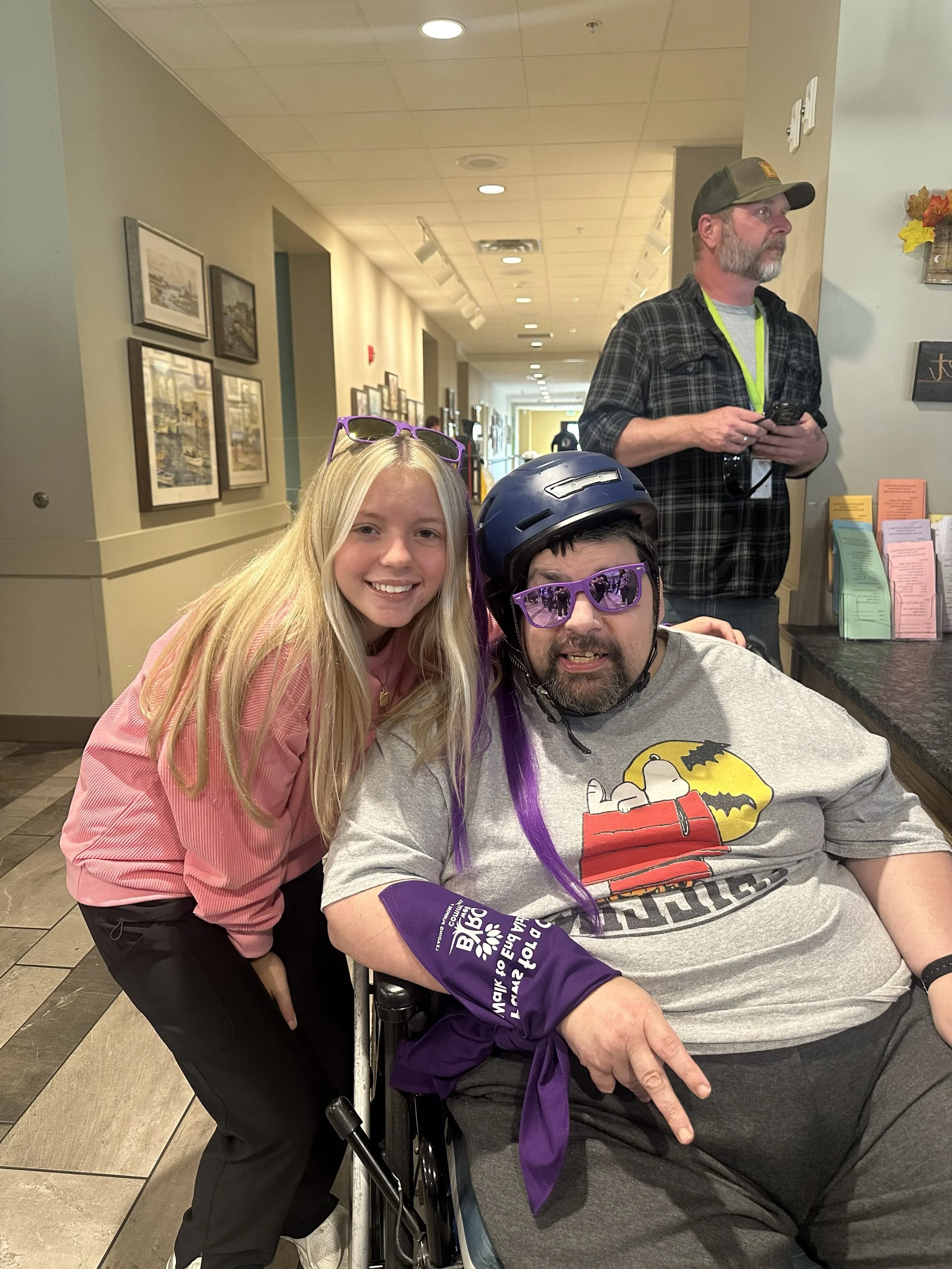 A young woman with long blonde hair wearing a pink outfit and sunglasses is smiling and leaning toward a man in a wheelchair. The man wears a helmet, purple sunglasses, a gray graphic T-shirt, and a purple sleeve on his arm. An older man stands in th