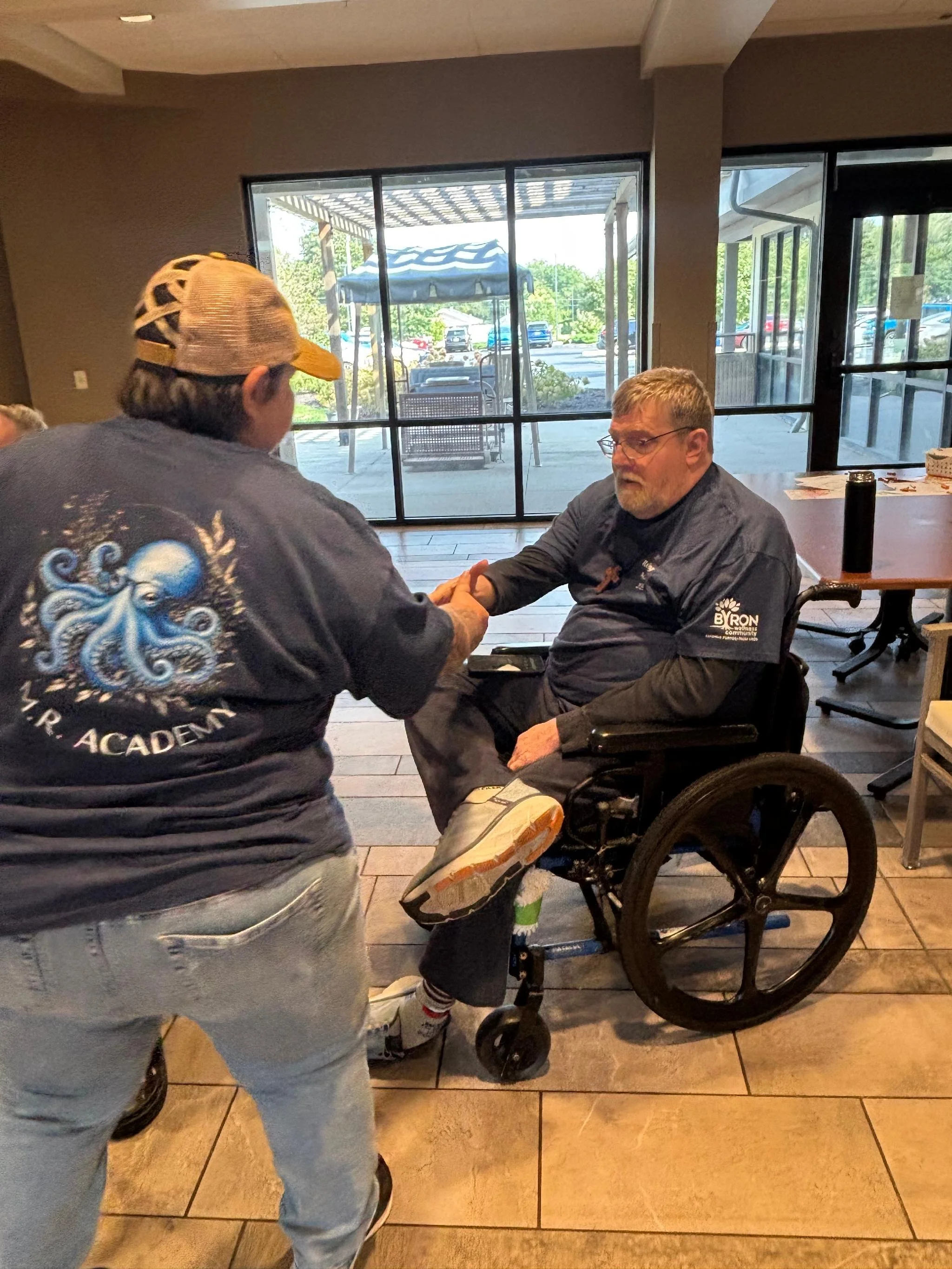 A man in a wheelchair holding hands with another person who is standing. They are inside a building with large windows, outside there is a patio with a canopy and parked cars.