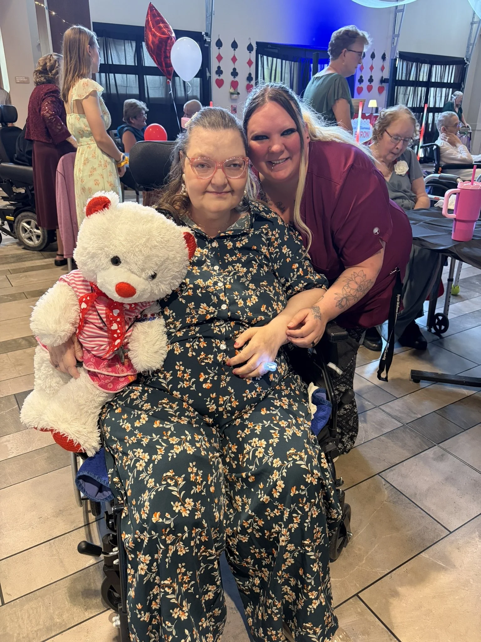 Two women sitting in wheelchairs smiling at the camera in a room decorated with balloons and playing cards theme, with other elderly people and caregivers in the background.