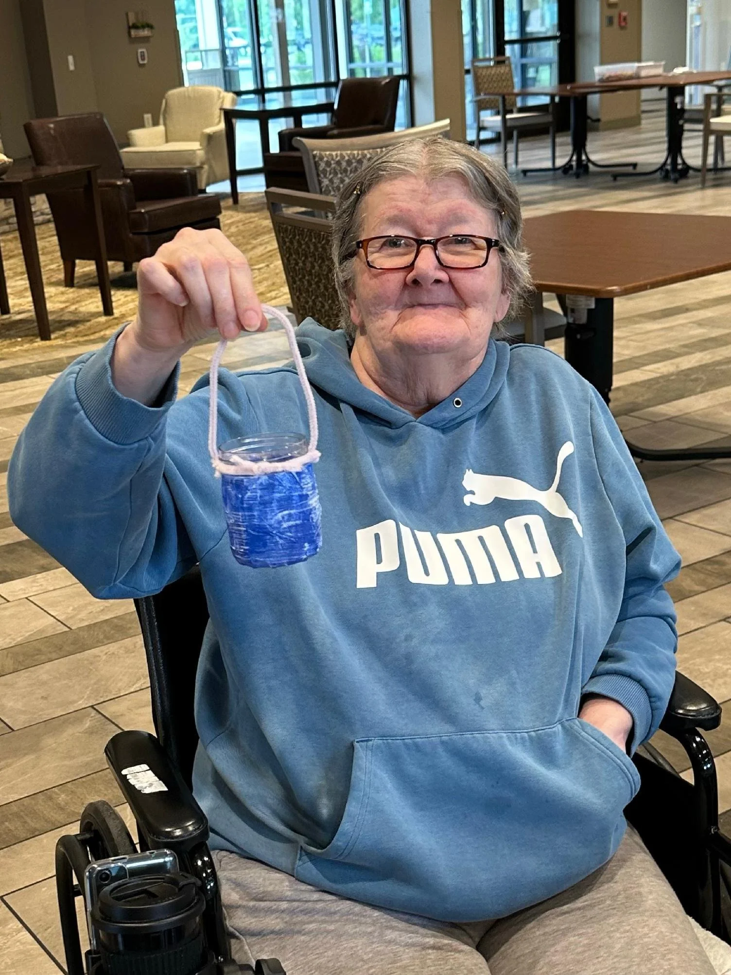 An elderly woman in glasses is sitting in a wheelchair inside a spacious room with large windows. She is holding a small, blue, and purple decorated face mask. She is wearing a blue Puma hoodie and gray pants, smiling slightly at the camera.