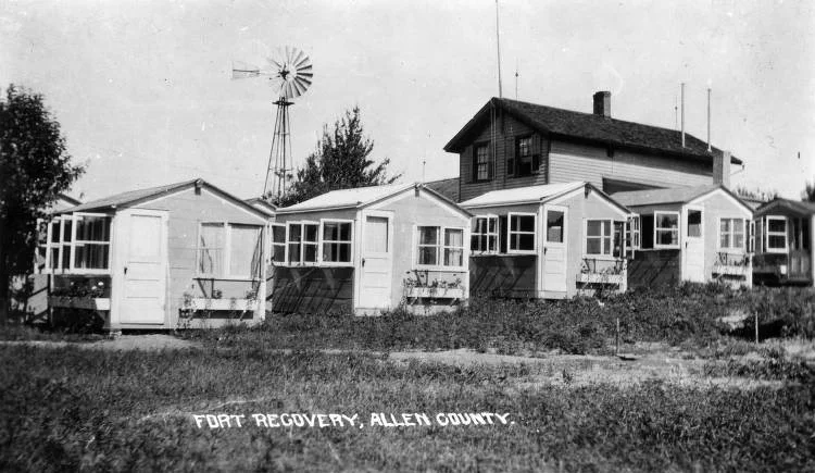 Black-and-white photo of a row of small houses with a larger house and windmill in the background, labeled "Fort Recovery, Allen County."