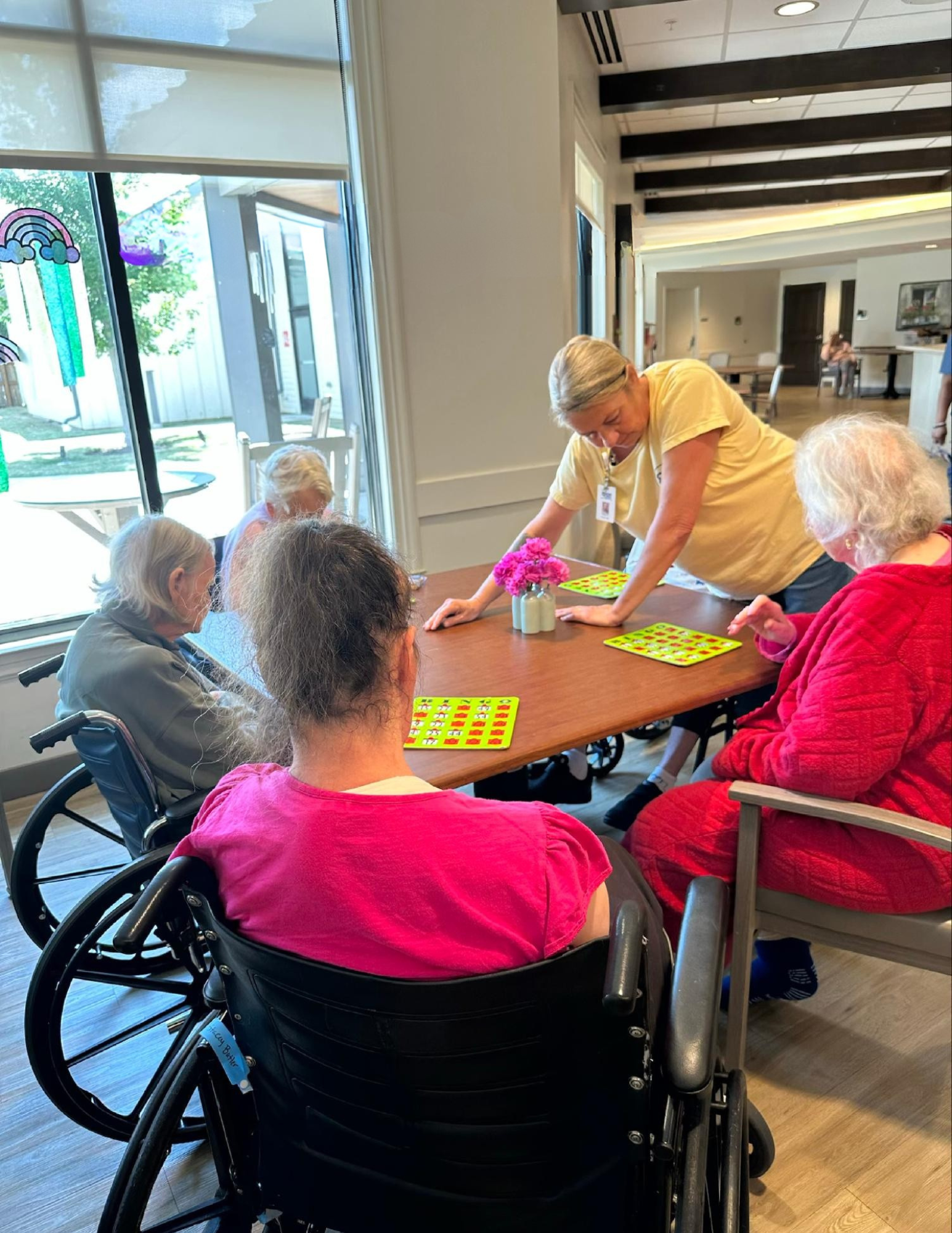 Group of elderly women playing bingo around a table with pink flowers in a vase, in a well-lit room with large windows and a view of an outdoor patio.