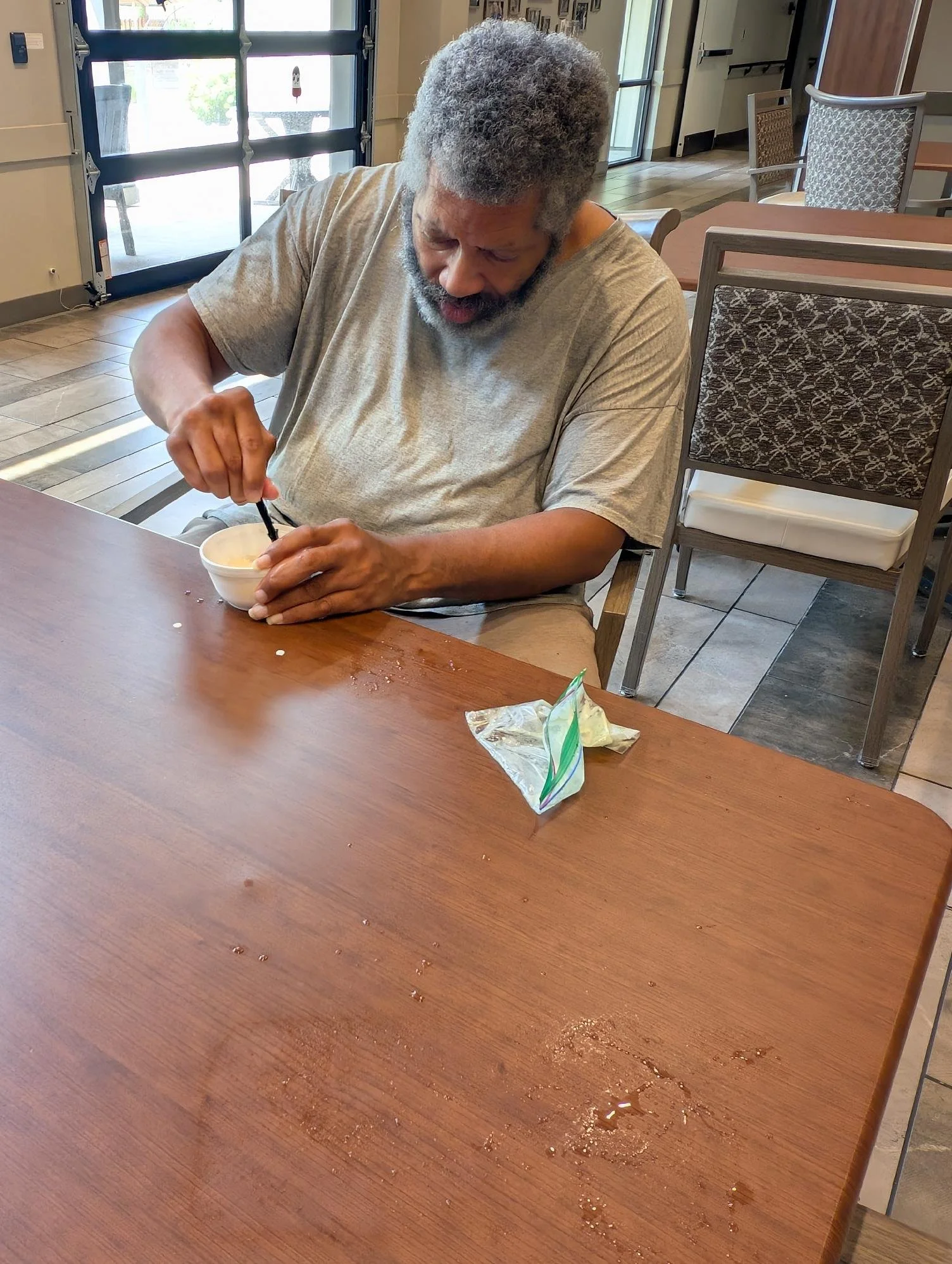 A senior man with gray hair and beard wearing a gray T-shirt sitting at a wooden table, using a spoon to eat from a bowl.