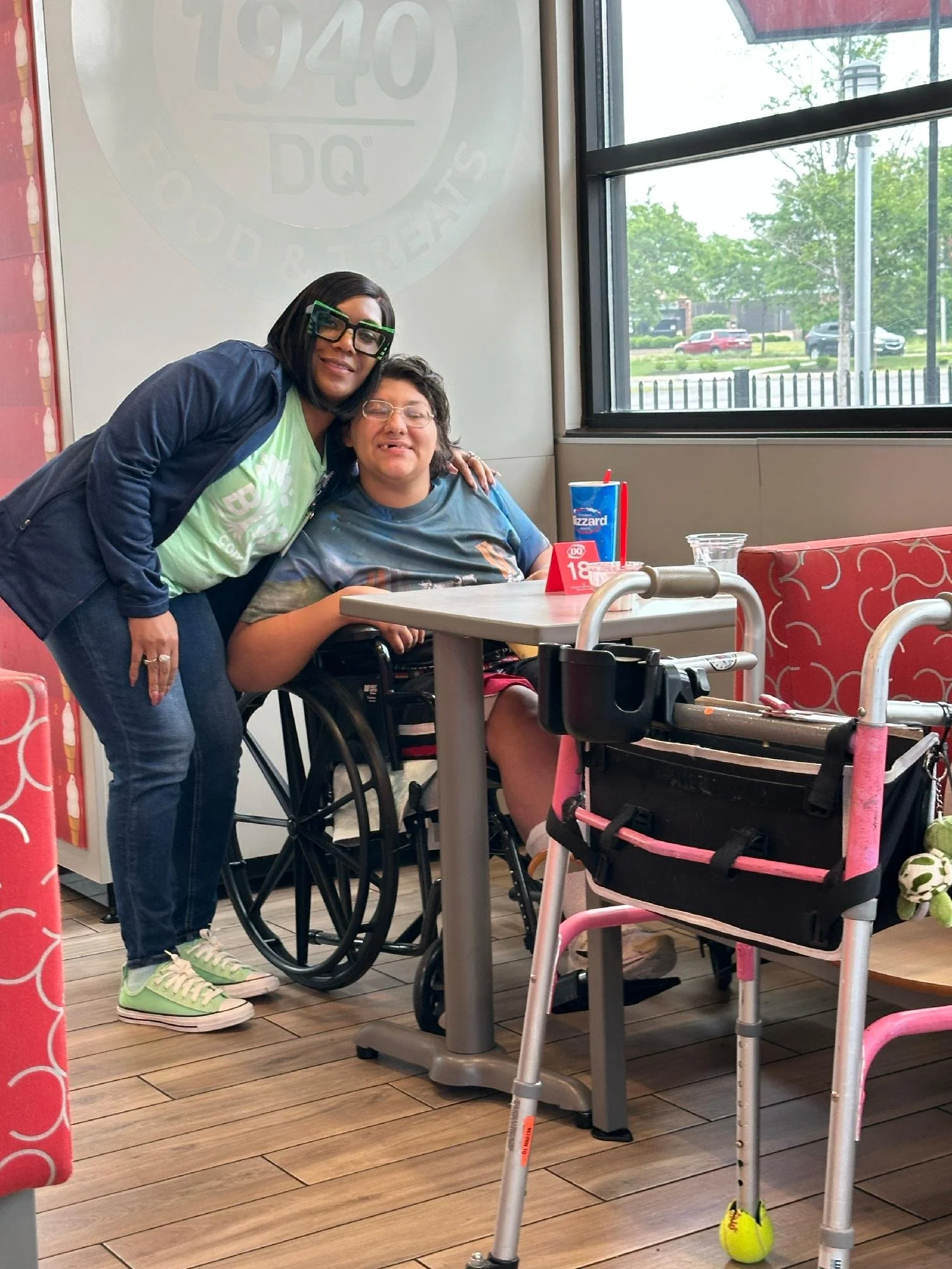 A woman in a wheelchair and another woman standing beside her inside a dairy restaurant, with large windows and a view of the parking lot and trees outside.