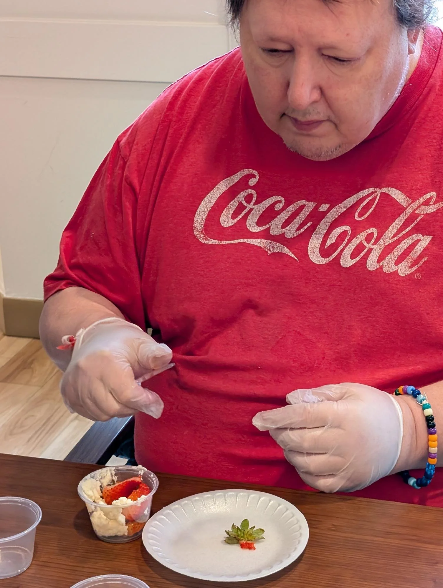 A person wearing a red Coca-Cola T-shirt and white gloves preparing strawberries and whipped cream in a small cup at a table with empty plates and cups.