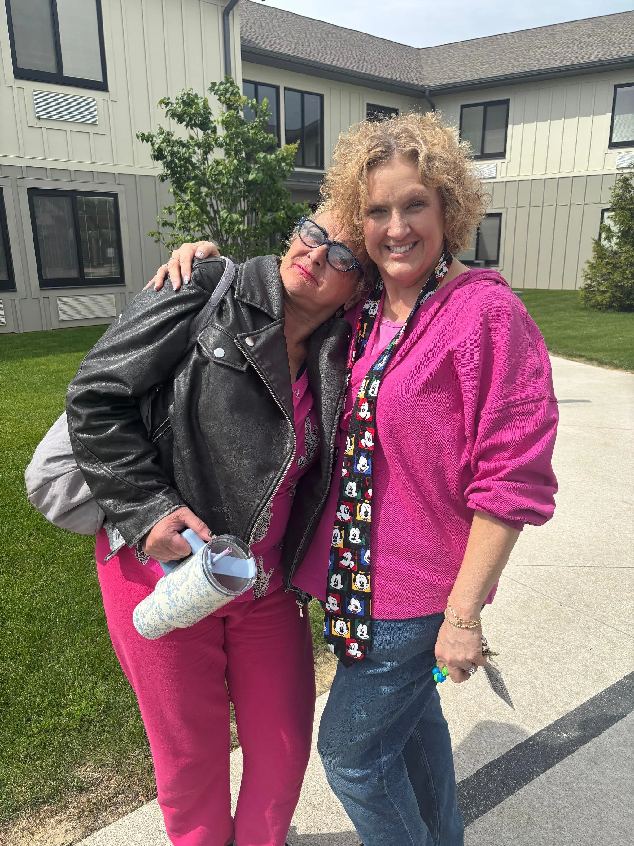 Two women standing close together, one with curly blonde hair and the other with short gray hair and glasses, smiling outdoors in front of a building with large windows and green grass.