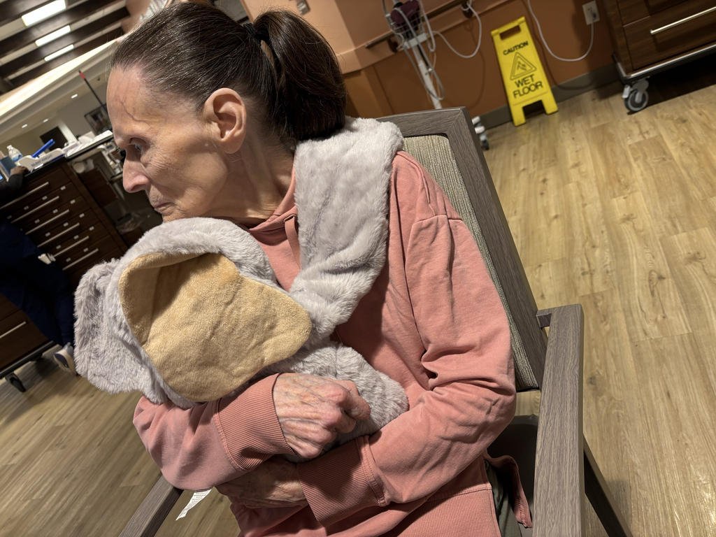An elderly woman sitting in a hospital waiting room, hugging a comforting stuffed animal. She has her eyes closed and appears to be resting or deep in thought. The room has wooden floors, medical equipment, and a yellow wet floor caution sign in the 