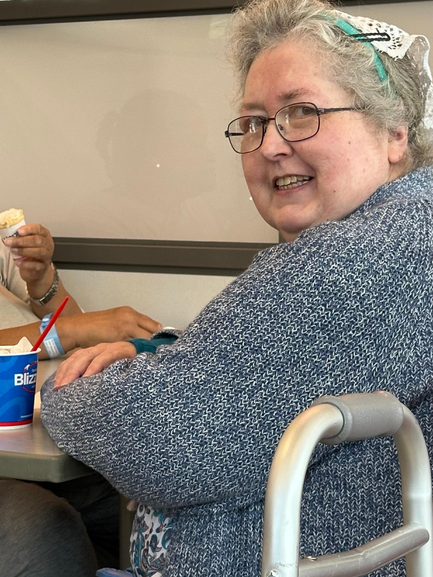 An elderly woman with gray curly hair and glasses smiling at the camera, sitting in a wheelchair at a table in a social setting.