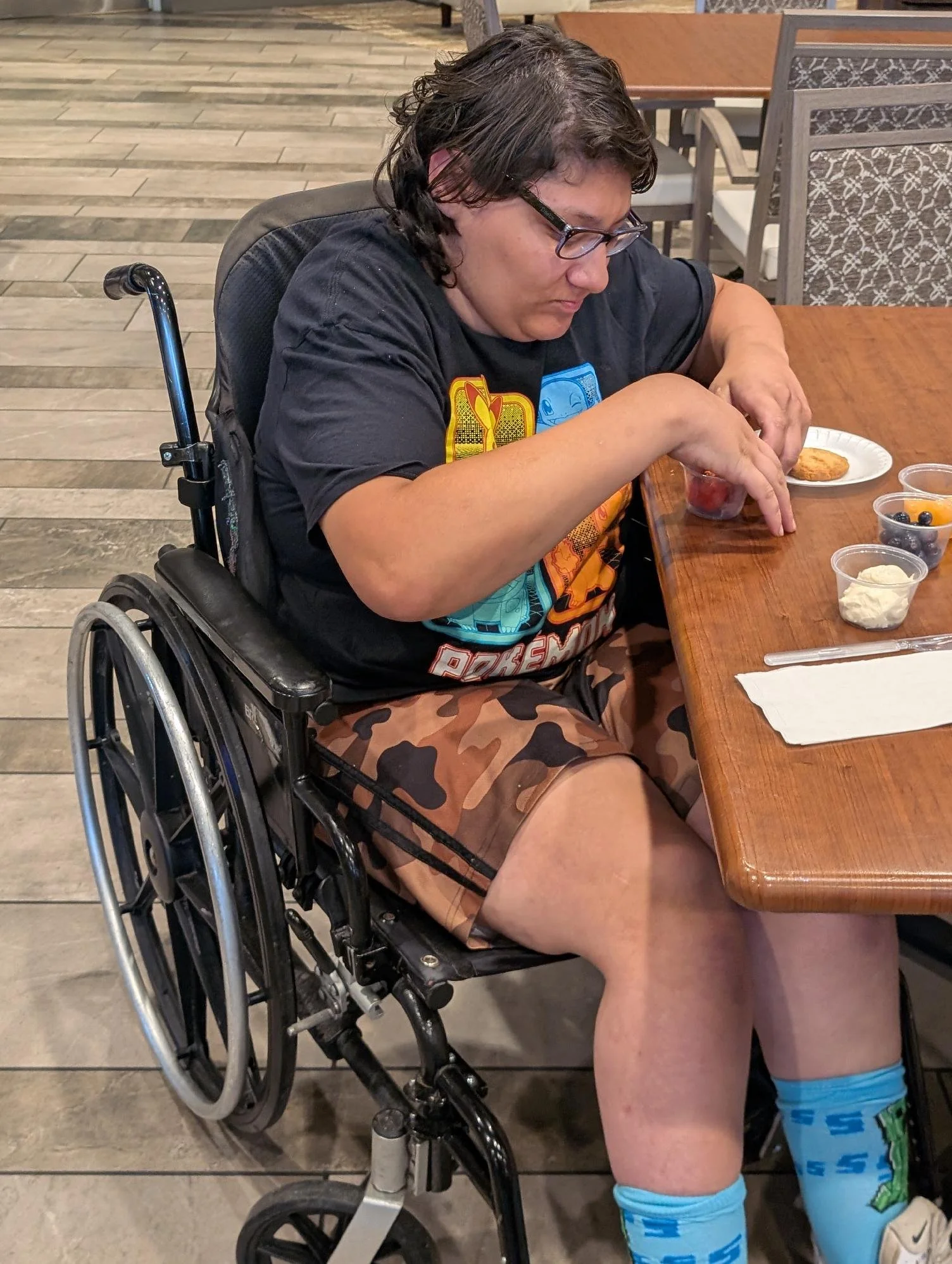 A man with glasses sitting in a wheelchair at a restaurant table, wearing a black t-shirt with colorful graphics, camouflage shorts, and blue cartoon character socks. He is reaching into a plastic cup with strawberries.