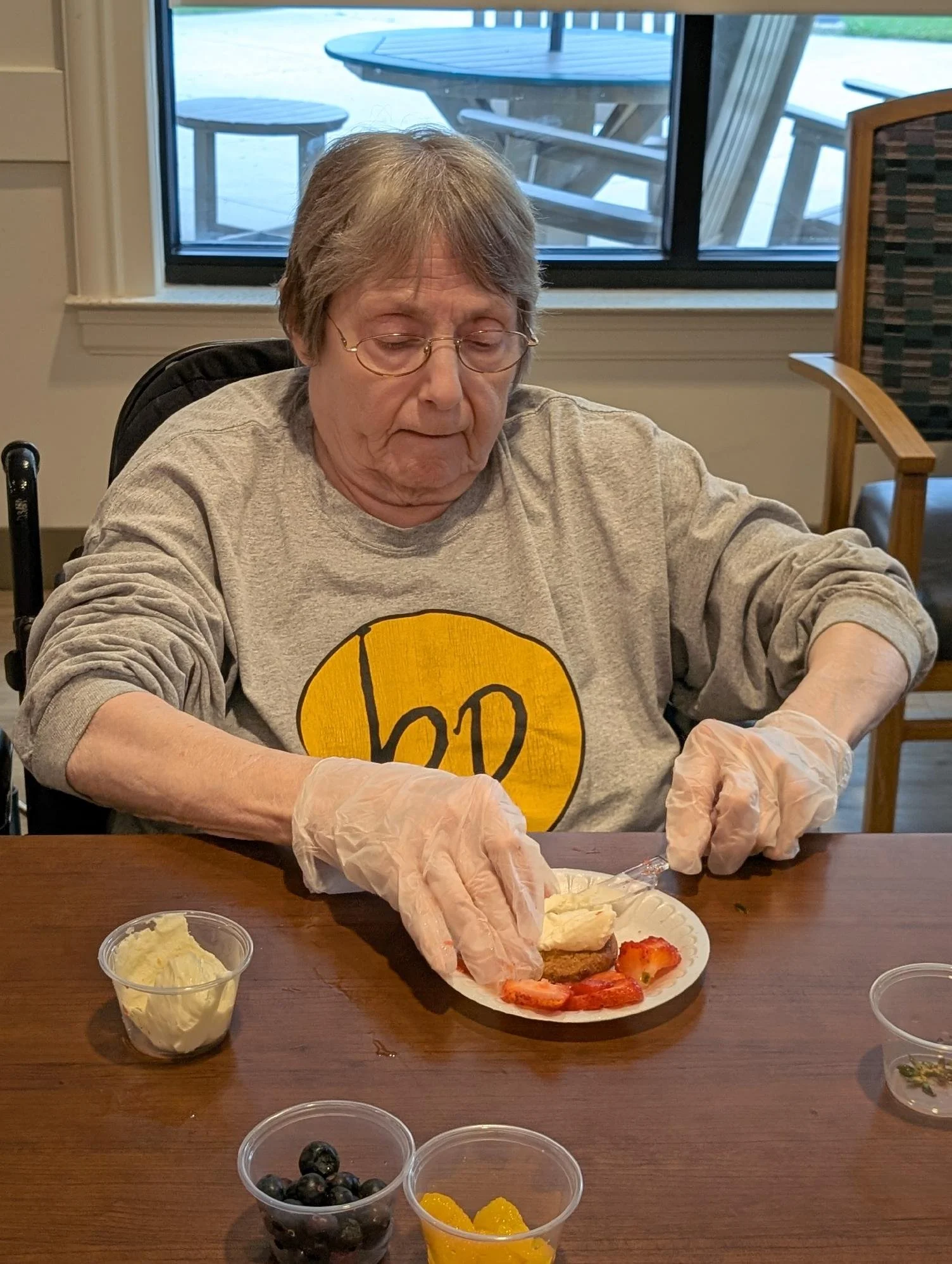 An elderly woman with glasses and gray hair sits at a table, wearing a gray sweatshirt with a yellow smiley face, preparing food with her hands in plastic gloves. There is a plate of strawberries and a piece of cake in front of her, along with small 
