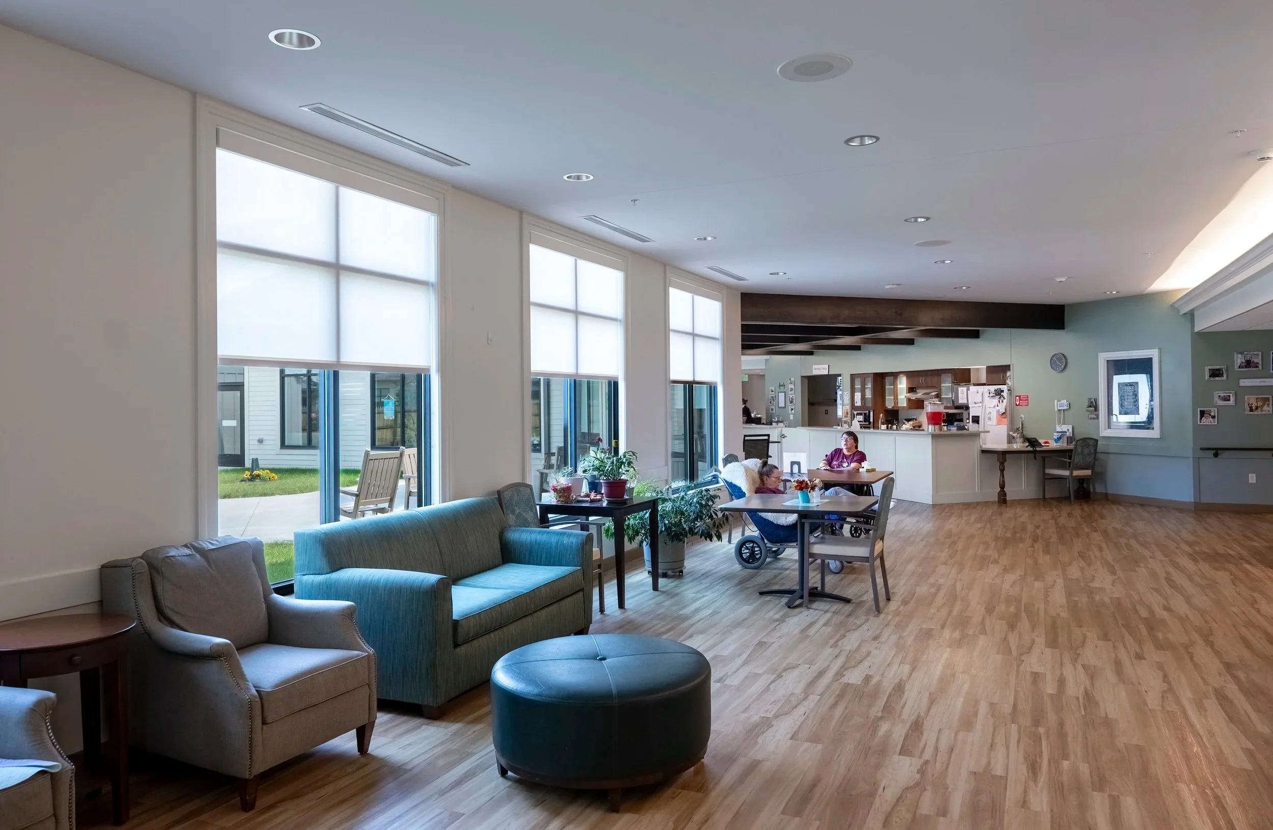 Interior of a community room with large windows, couches, chairs, and a table where two people are seated. The background includes a kitchen area and decorative photo frames on the wall.