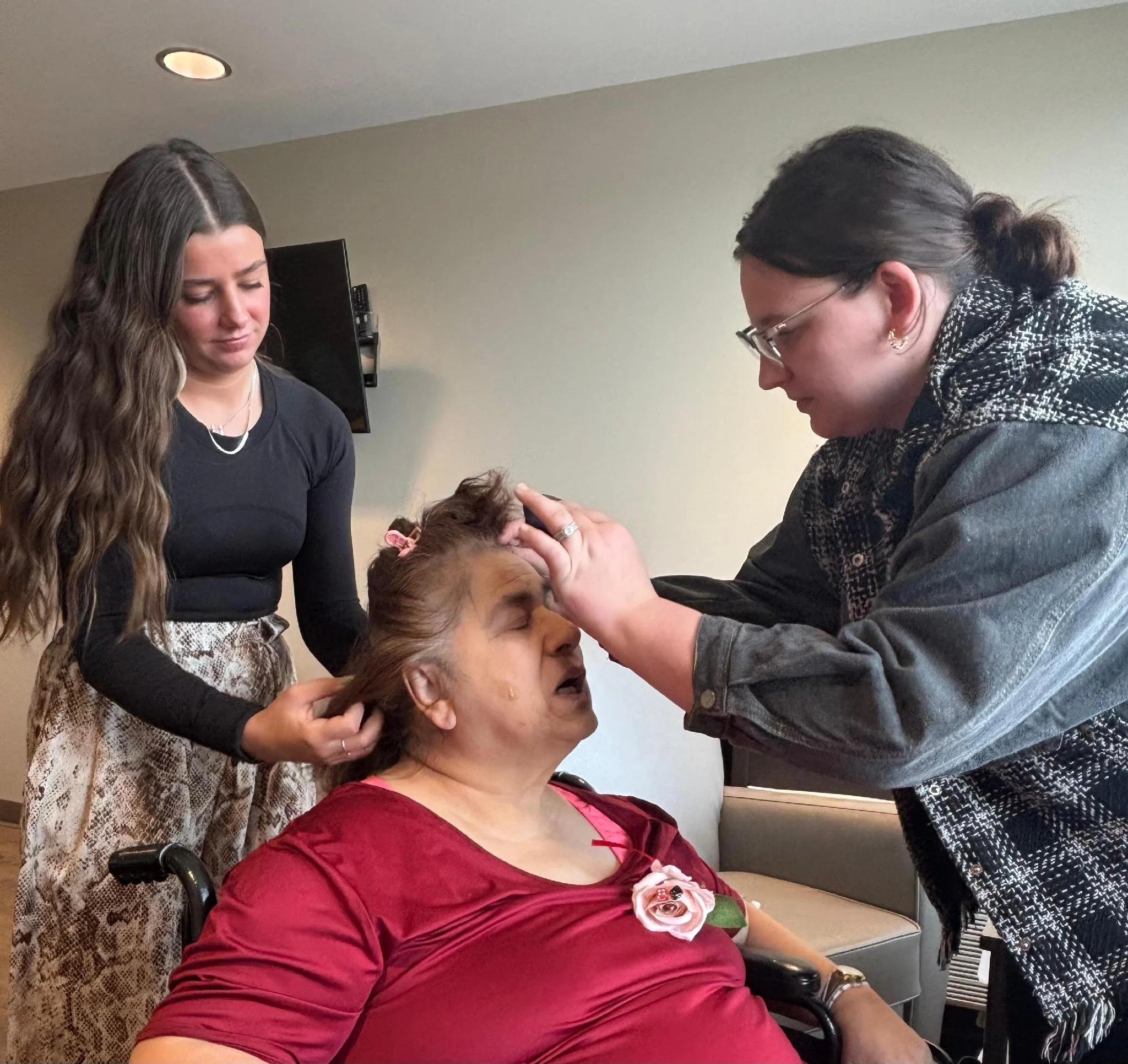 A woman in a maroon dress with a pink flower brooch is sitting in a wheelchair. A woman with glasses and dark hair tied in a bun, wearing a black and white patterned jacket, is leaning forward and holding the woman's forehead. Another woman with long