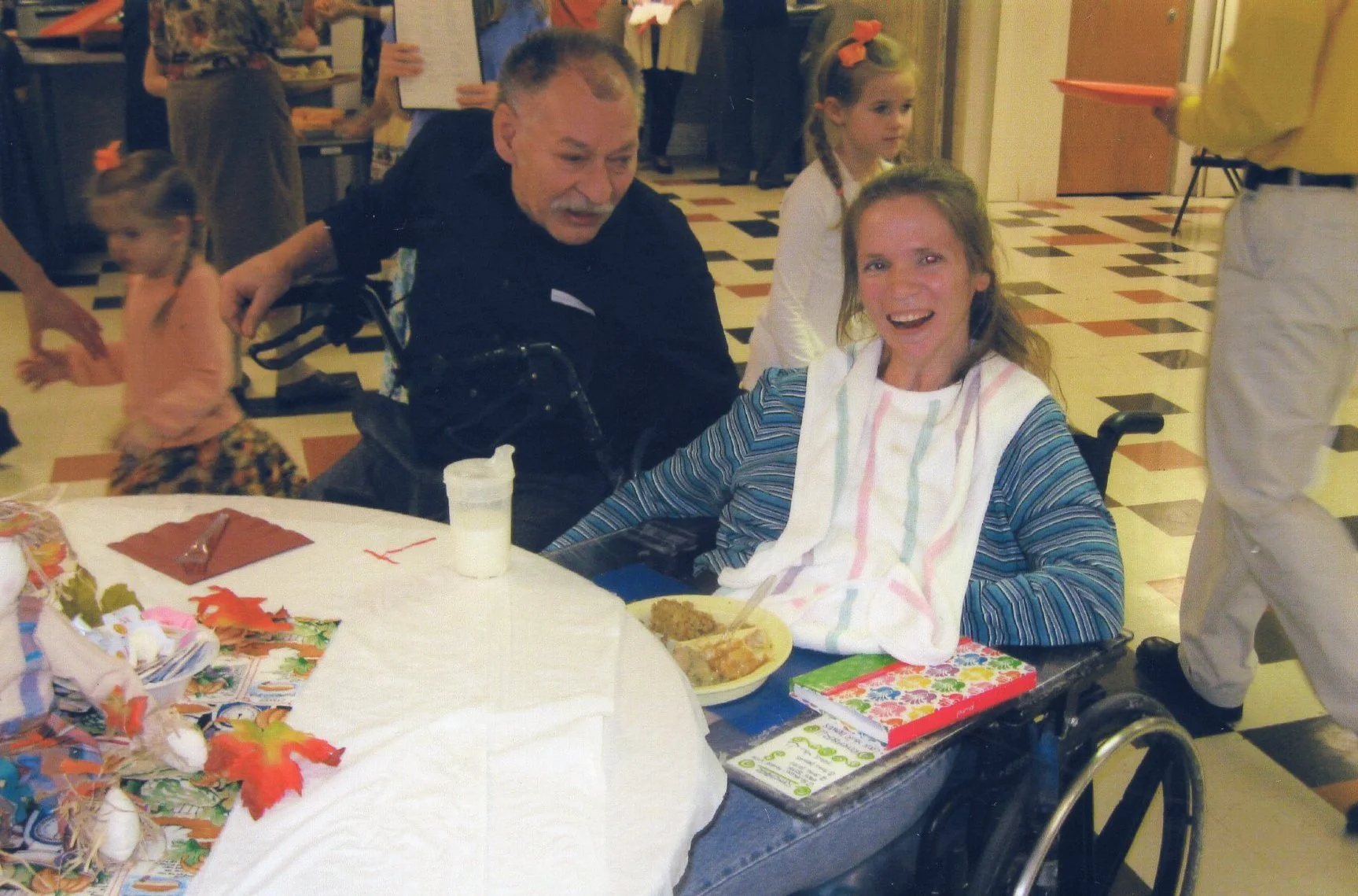 A woman in a wheelchair with a bib around her neck smiling at a man and young girl at a party table, with children and adults in the background.