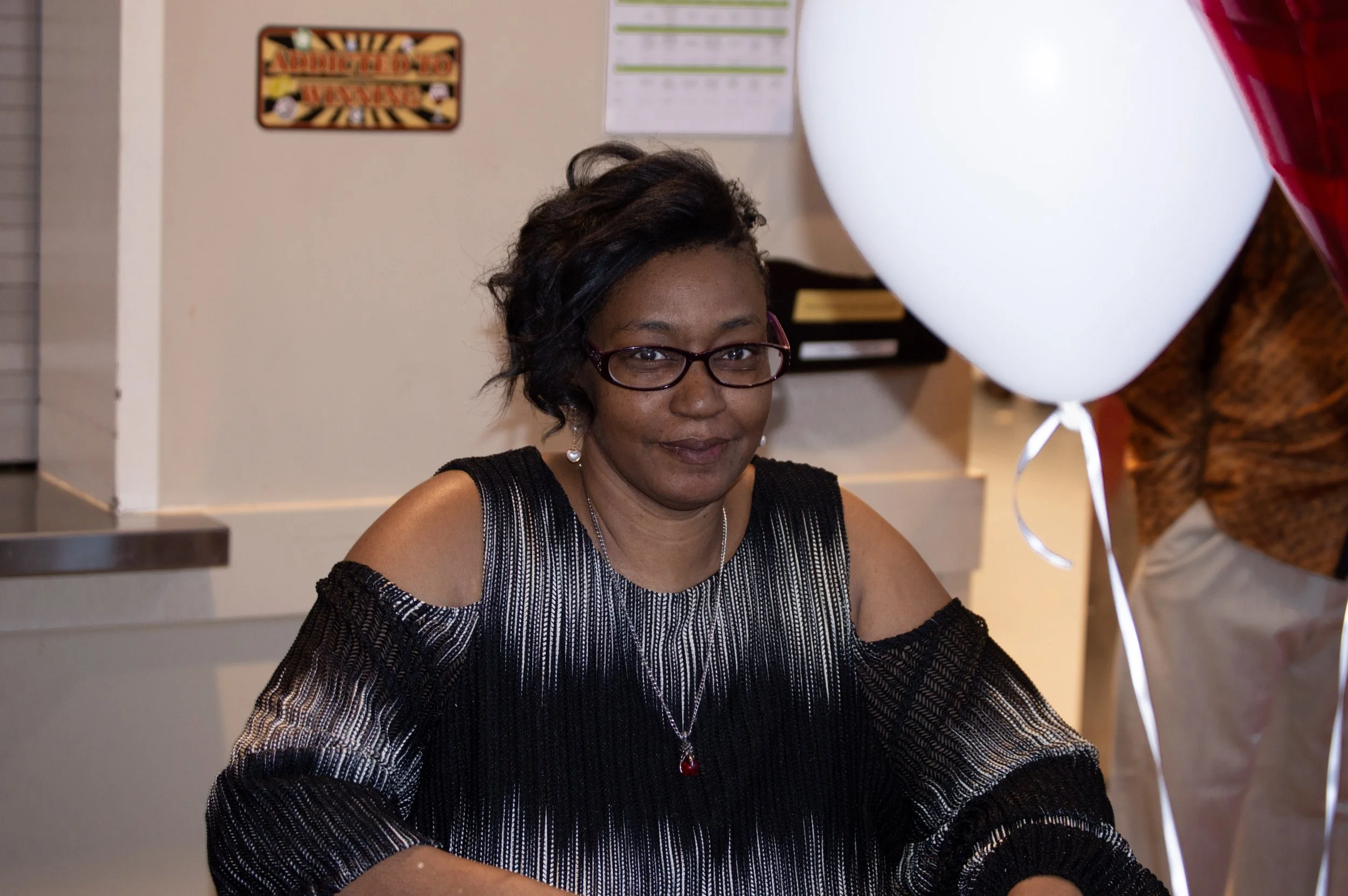 A woman with dark hair, glasses, and earrings sitting at a table, smiling at the camera, with a large white balloon nearby and a person in patterned clothing partially in view.