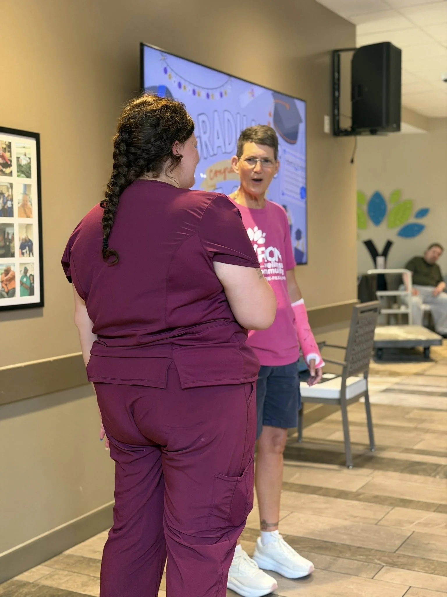 Two women talking in a restaurant or cafe setting. The woman in the foreground has dark curly hair tied in a braid and is wearing a maroon scrub top and pants. The woman in the background is wearing glasses, a pink t-shirt, and jeans, with white snea