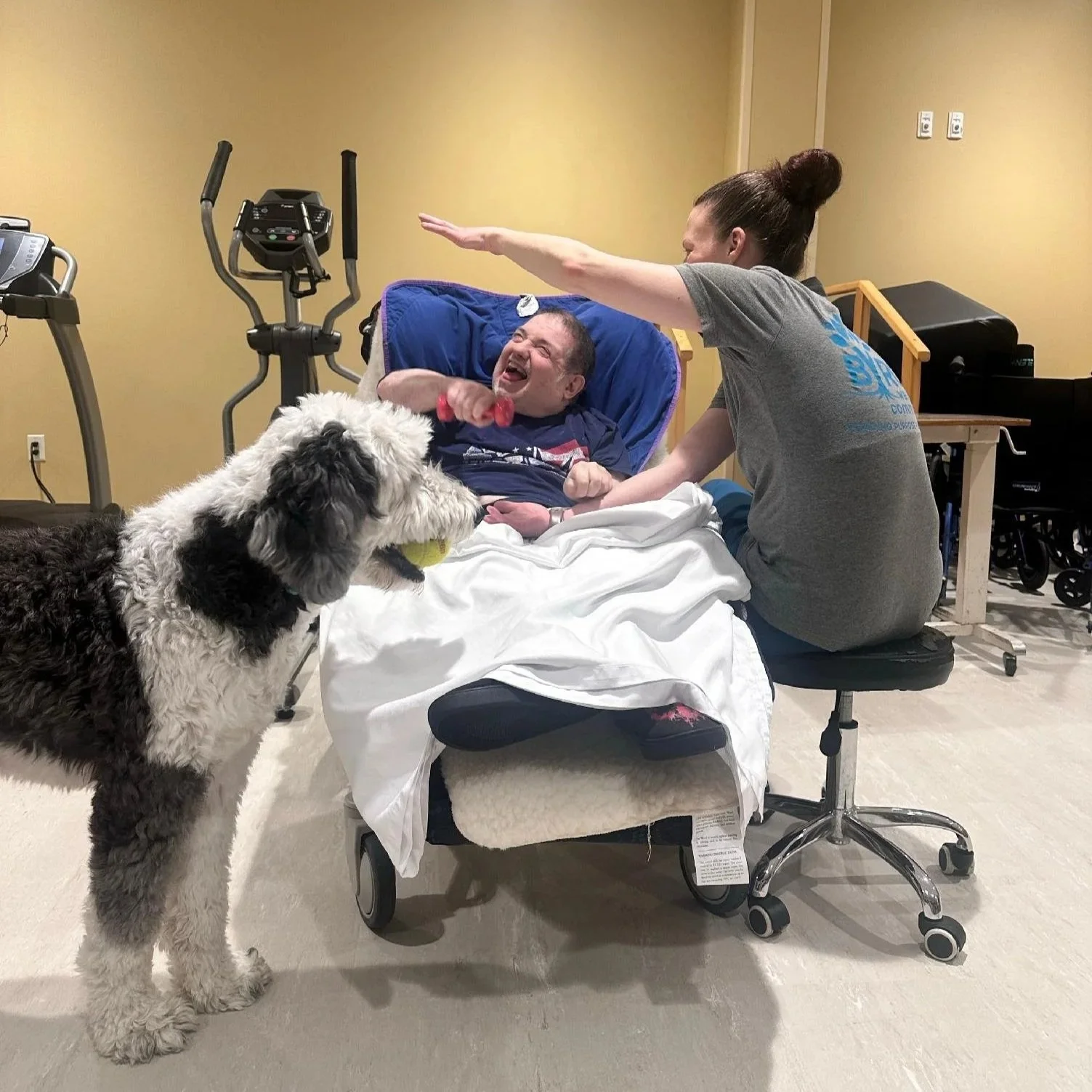 A person with a dog in a hospital room, the person is holding a pink dumbbell and grinning, while the dog holds a tennis ball in its mouth, and a caregiver is sitting near the patient with her arm extended.