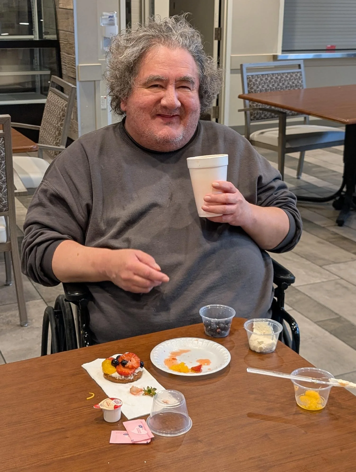 A man with gray, curly hair and a gray sweatshirt sits in a wheelchair at a wooden table, holding a white foam cup. The table has leftover dessert plates and snack containers, some with food and some empty. The background shows a room with chairs, ta