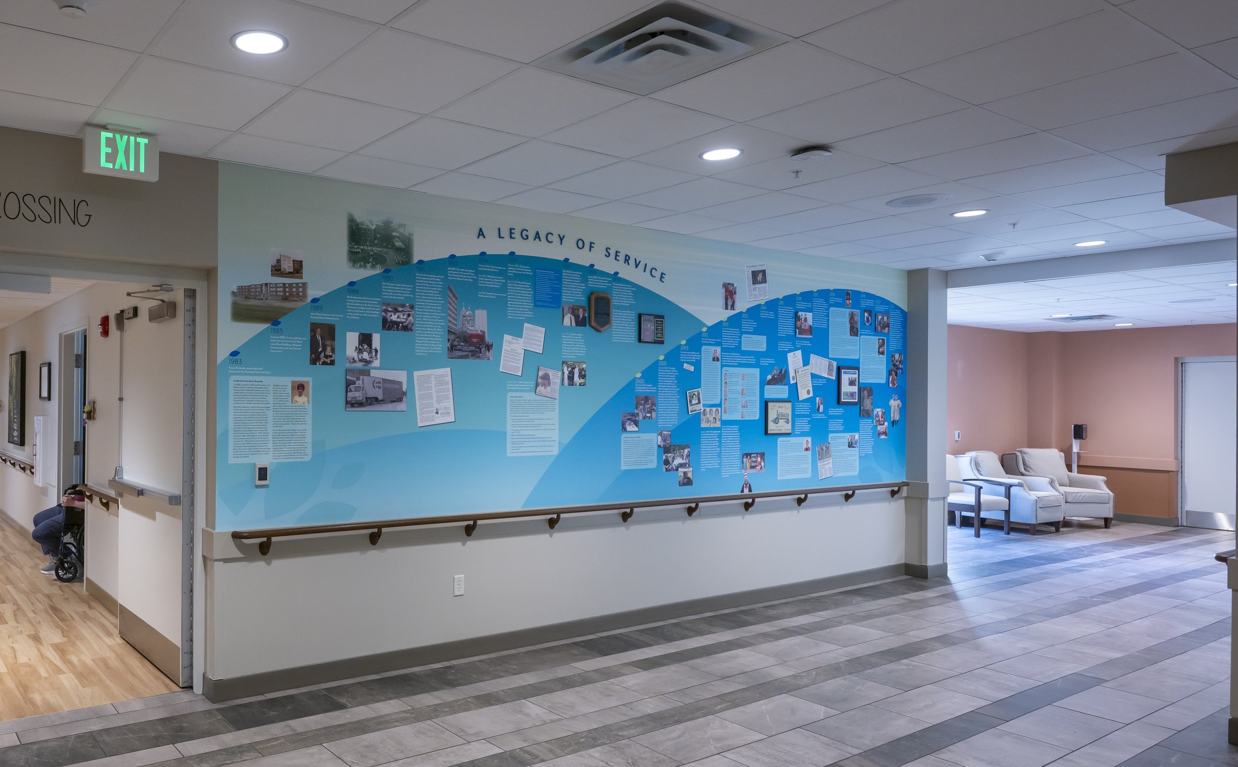 Hospital corridor with a blue informational wall display titled "A Legacy of Service," featuring photos and text about historical events. To the left, a wheelchair is visible near a door. To the right, there are seating areas with chairs and a hallway leading to more rooms. The ceiling has recessed lighting, and there is an illuminated EXIT sign above a door.
