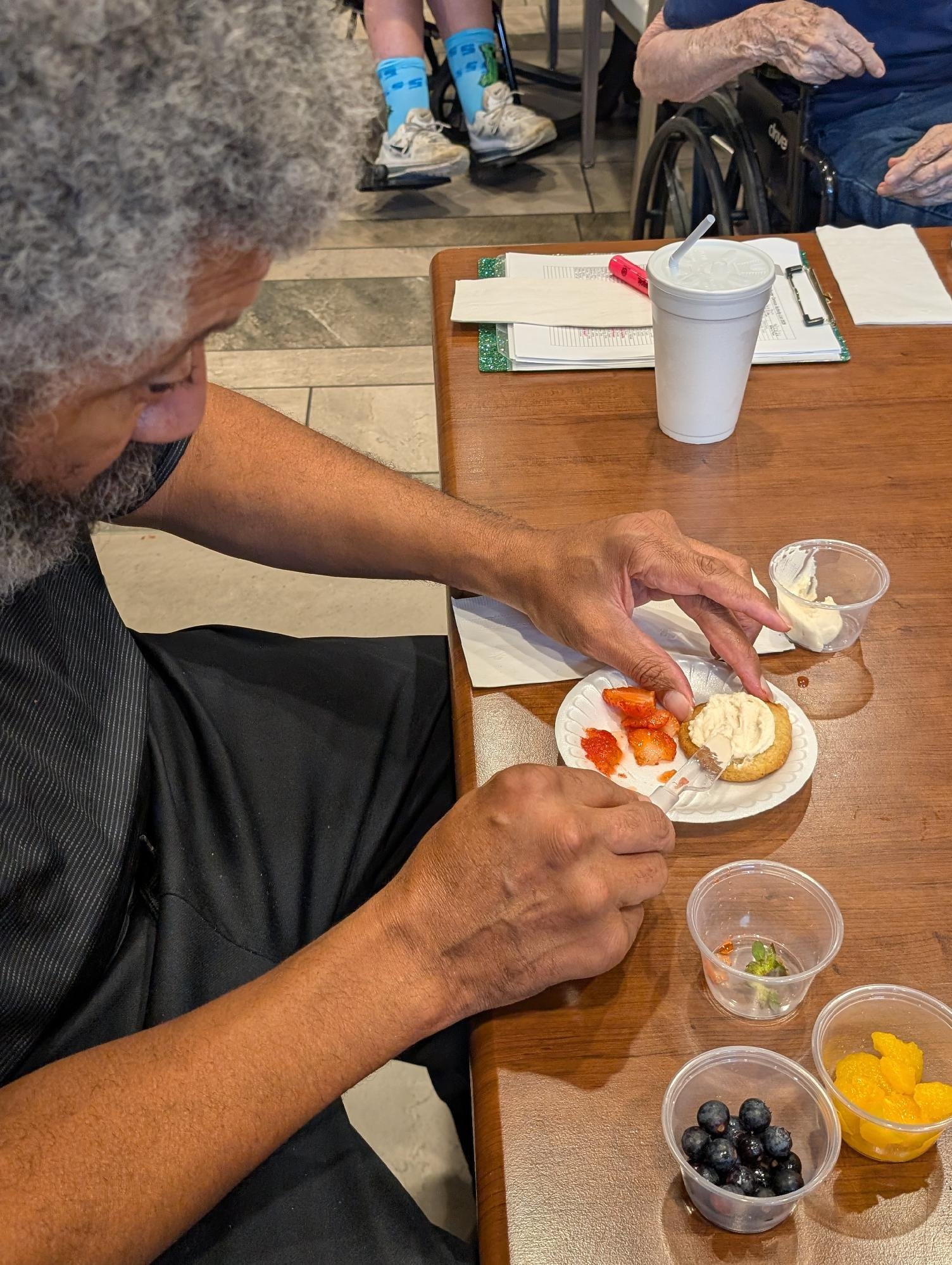 An elderly man with gray curly hair and a beard is sitting at a wooden table, eating a dessert with strawberries and whipped cream. The table also has a white cup with a straw, a small plastic container of ice cream, and several bowls of berries and 