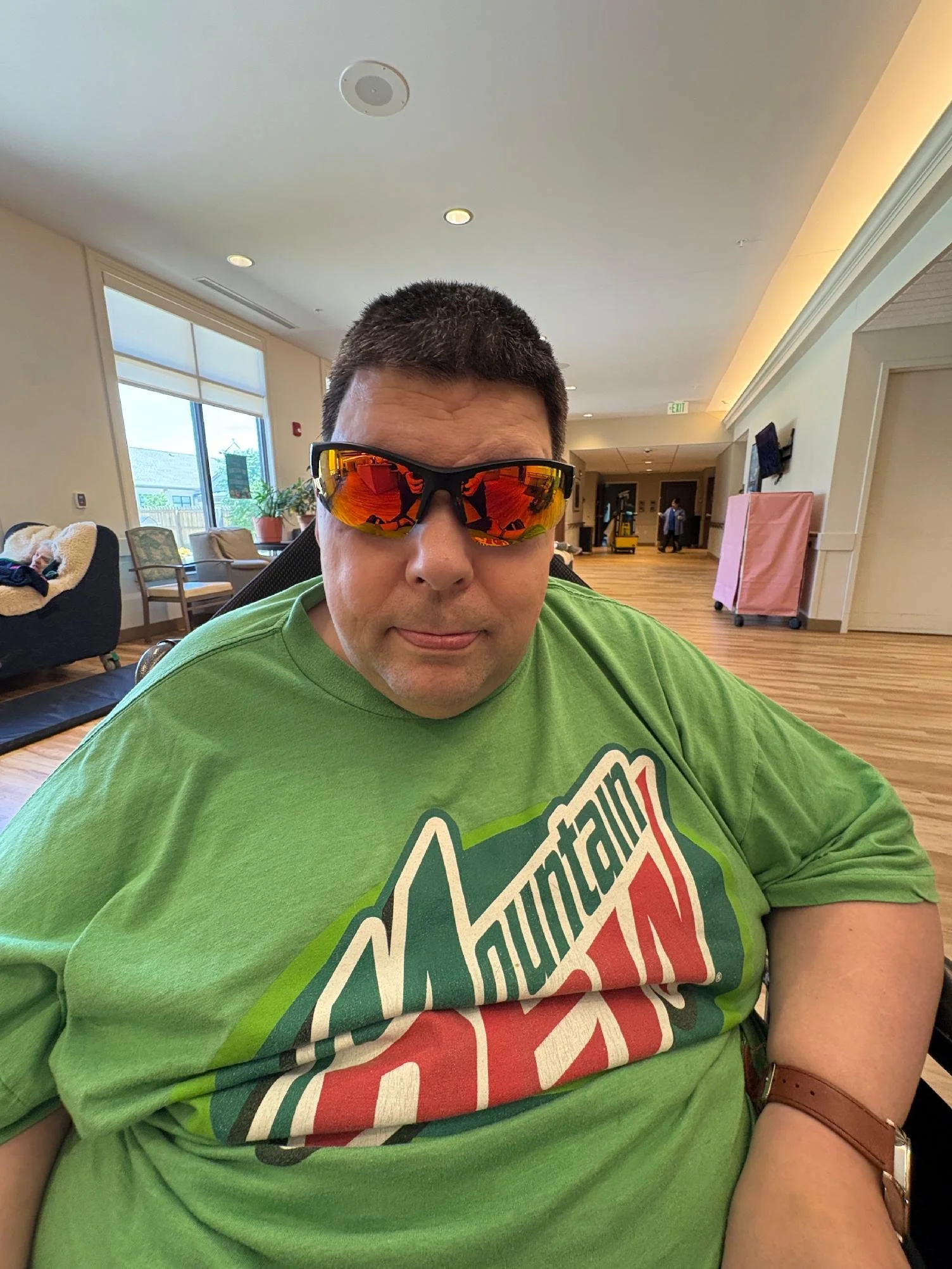 A man wearing reflective sunglasses and a bright green T-shirt with a partially visible logo, sitting indoors in a lounge area with wooden flooring, beige walls, and large windows.