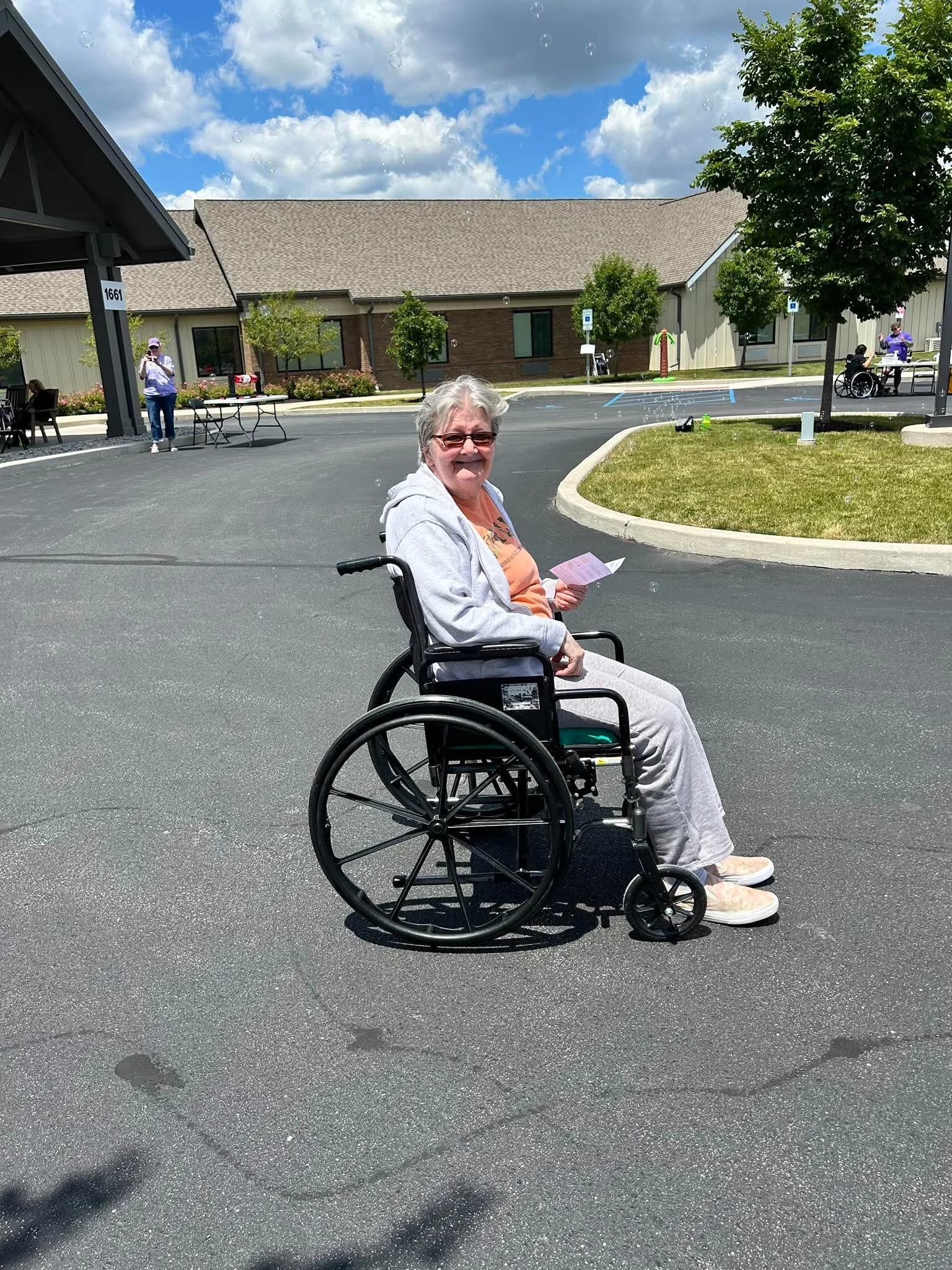 Smiling elderly woman in glasses sitting in a wheelchair outdoors on a sunny day, holding a piece of paper, with several trees, a building, and people in the background.