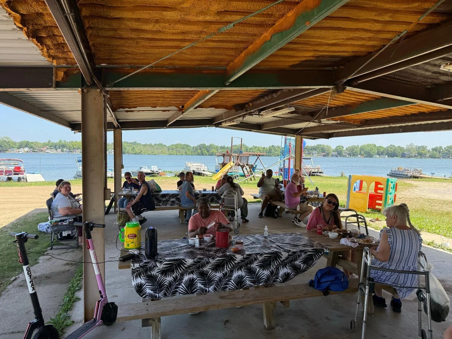 Group of people having a meal under a pavilion near a lake with boats and docks in the background, with children’s play equipment nearby.