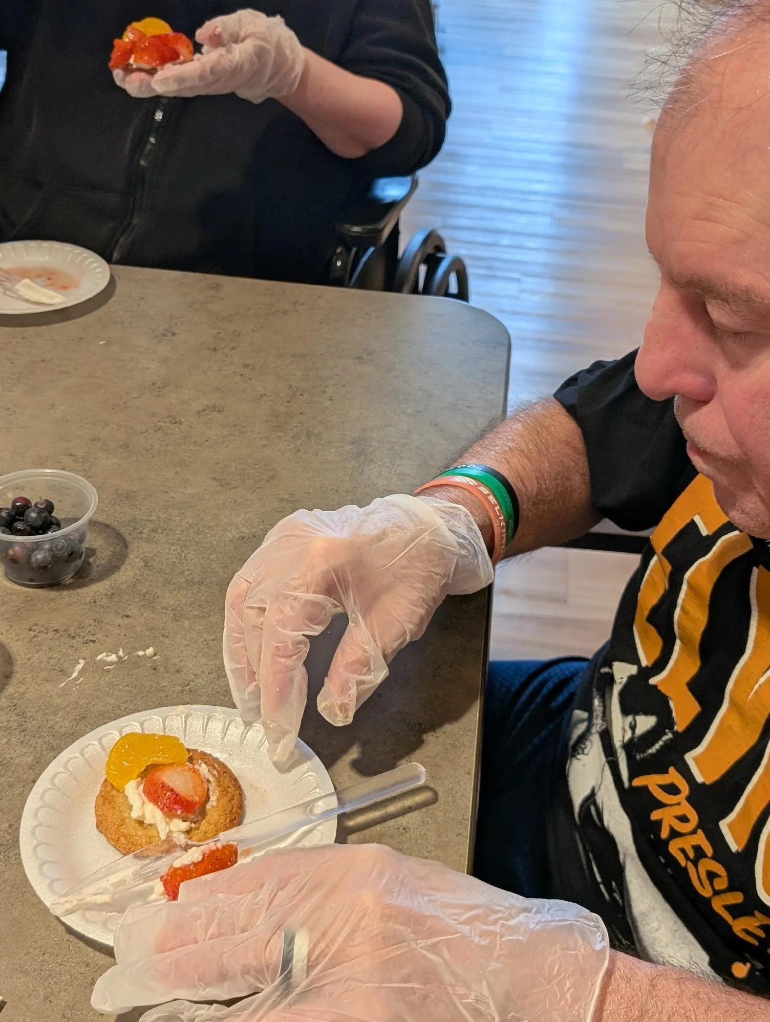 Person with a watch and bracelets preparing a fruit-topped dessert or pastry with strawberries, peaches, and whipped cream on a paper plate, at a table with blueberries and a fork.