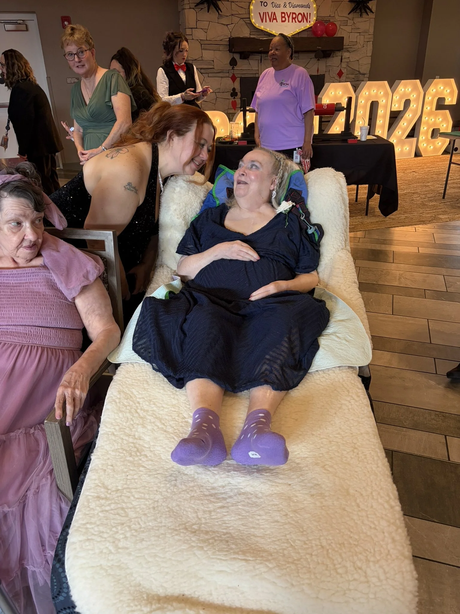 A woman with red hair leaning over and smiling at an elderly woman lying in bed with purple socks, in a room decorated for a celebration with large illuminated numbers and a sign that reads "Viva Byron!" in the background.