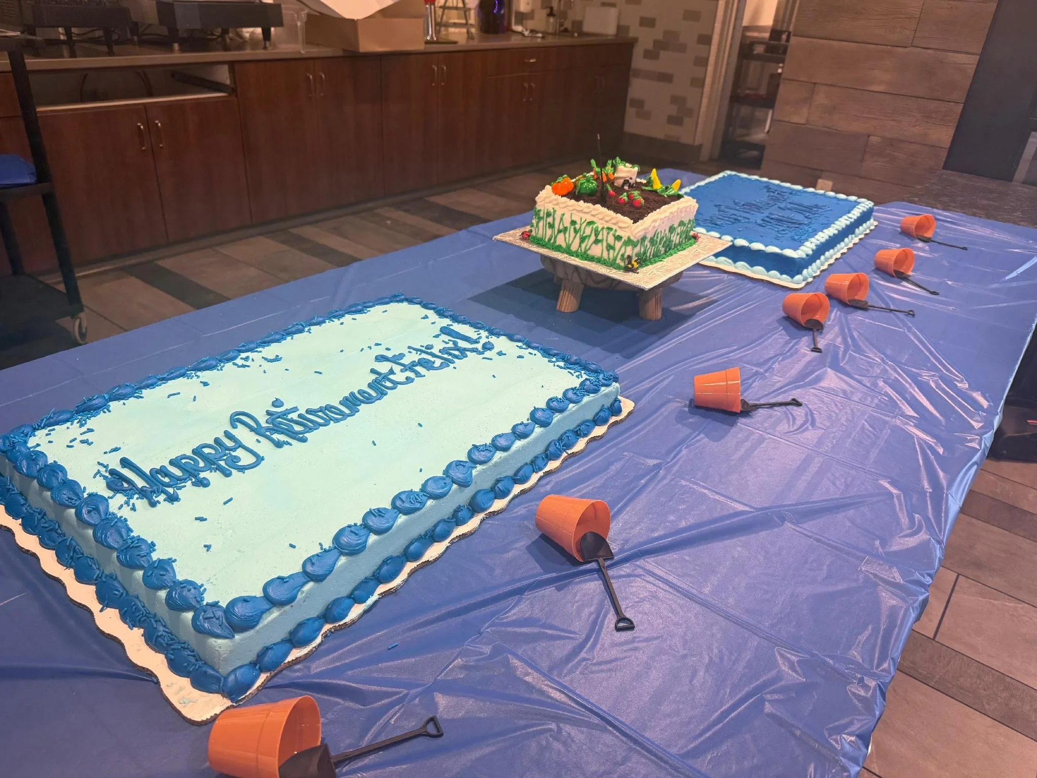 A decorated celebration cake on a table with blue tablecloth, surrounded by orange cups and utensils, in a room with wood and brick interior walls.