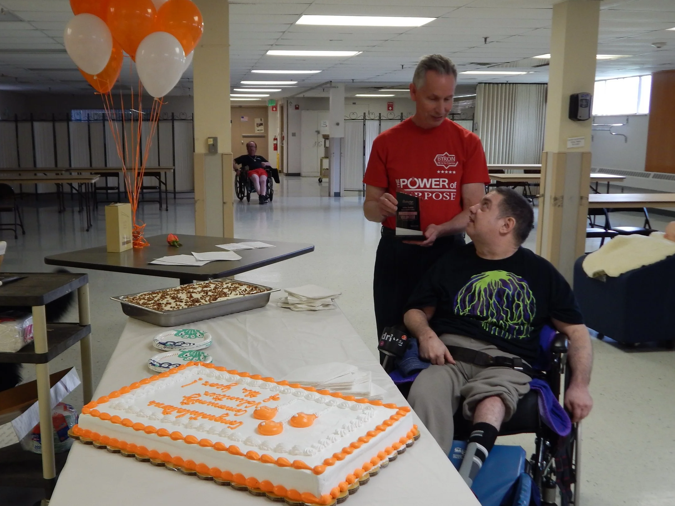 A celebration with a large rectangular white cake decorated with orange piping, surrounded by smaller treats, in a community hall with balloons and people, including a man in a wheelchair and another man standing.