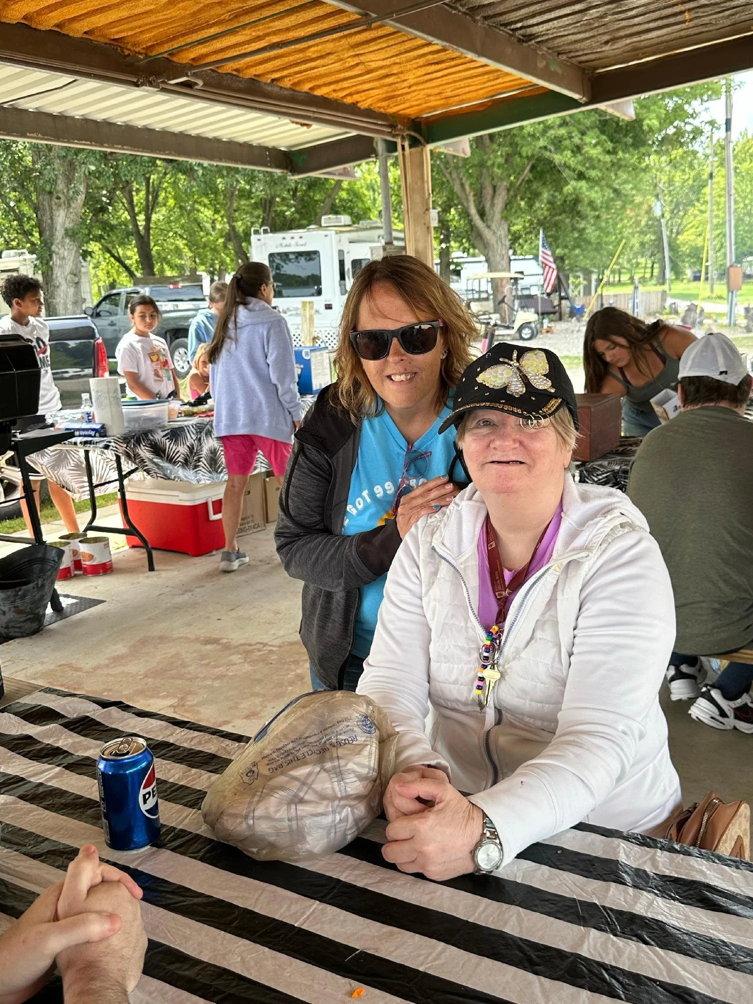 Two women sitting at a table under a canopy, smiling for a photo. The woman in front is wearing a white jacket and a black cap with butterfly design, while the woman behind her has sunglasses and a blue shirt. In the background, there are people, tab