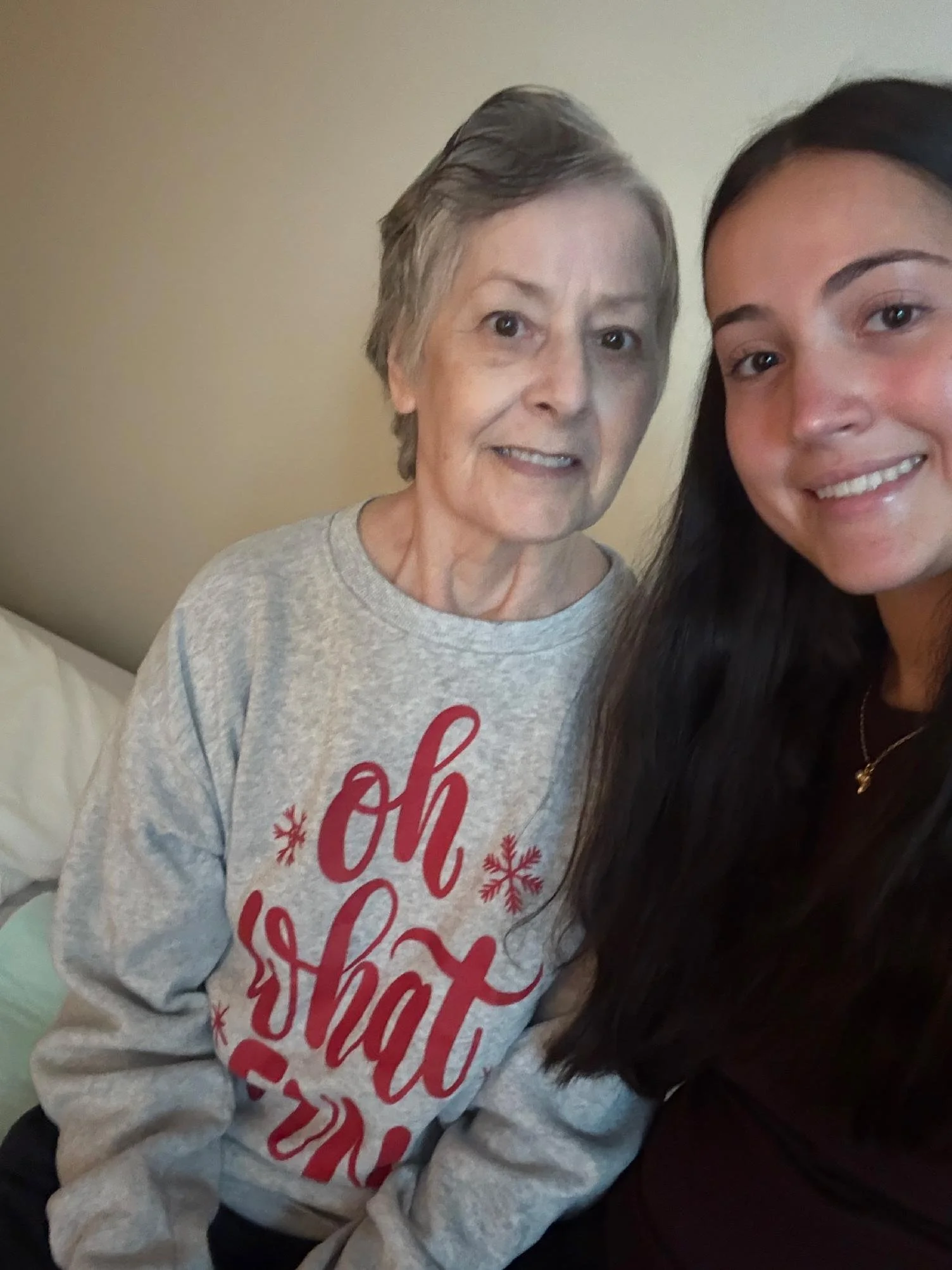 Close-up of two smiling women, one elderly with short gray hair wearing a gray holiday sweatshirt, and a younger woman with long dark hair, in a room with beige walls.