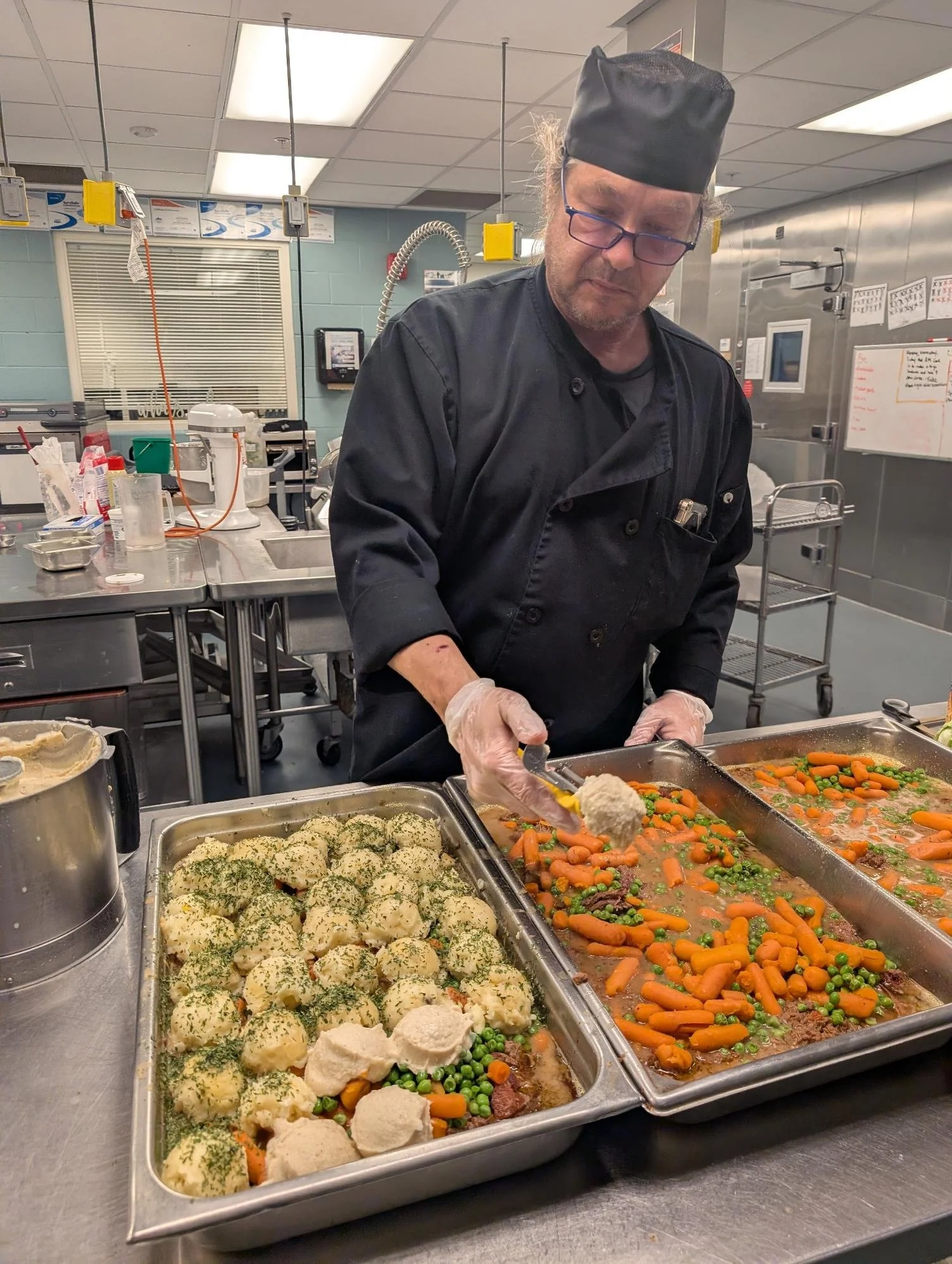 A chef in a commercial kitchen preparing food. There are trays of cooked vegetables and mashed potatoes with parsley on a stainless steel counter.