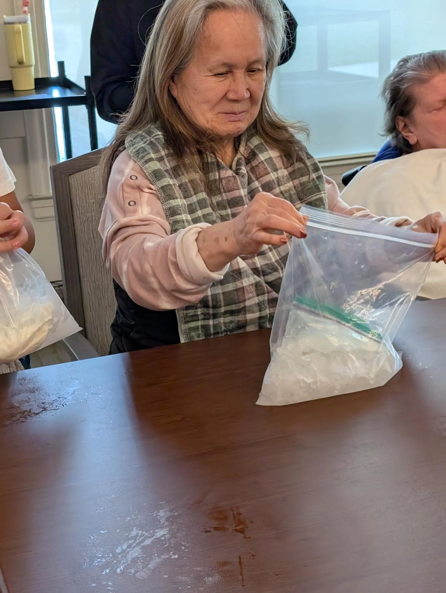 An elderly woman with long gray hair and a plaid vest over a pink shirt, holding a plastic bag with her hands, sitting at a table during daytime.