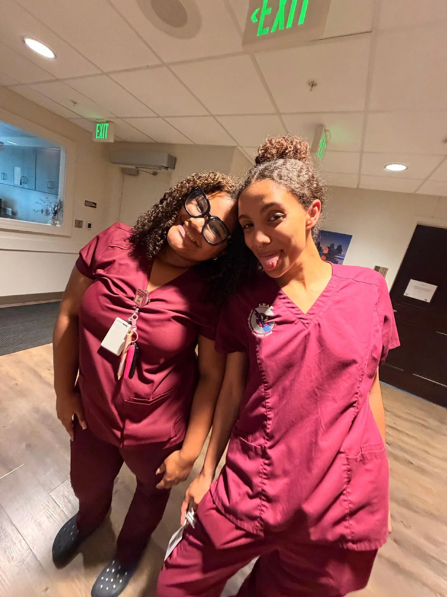 Two individuals wearing matching burgundy scrubs, standing closely and smiling playfully, with one sticking her tongue out, in an indoor setting with ceiling lights and emergency exit signs.