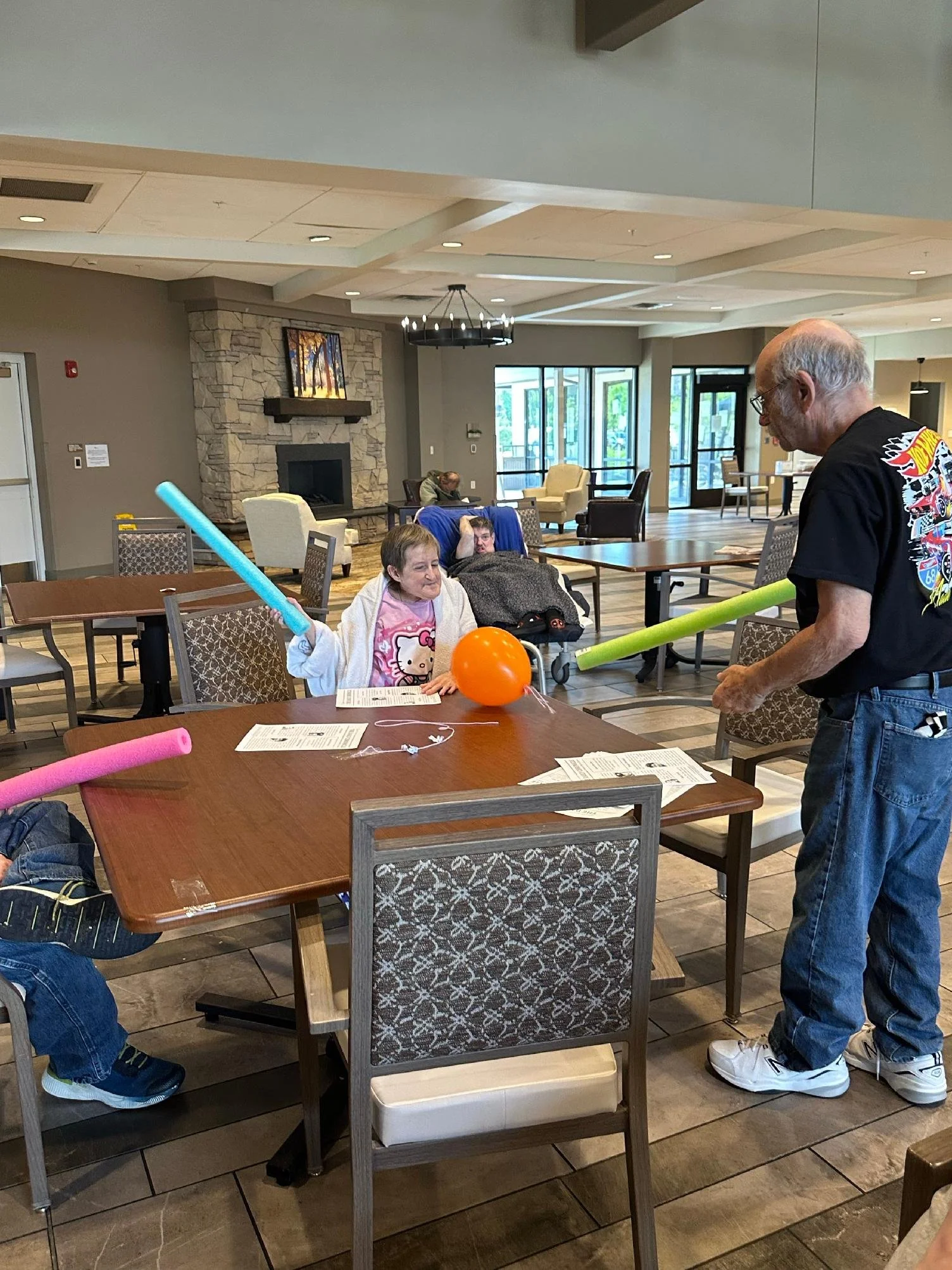 A group of people in a dining area with tables and chairs. One person is holding a foam noodle, and a child at the table is smiling. There is a fireplace with a painting above it, and large windows letting in natural light.
