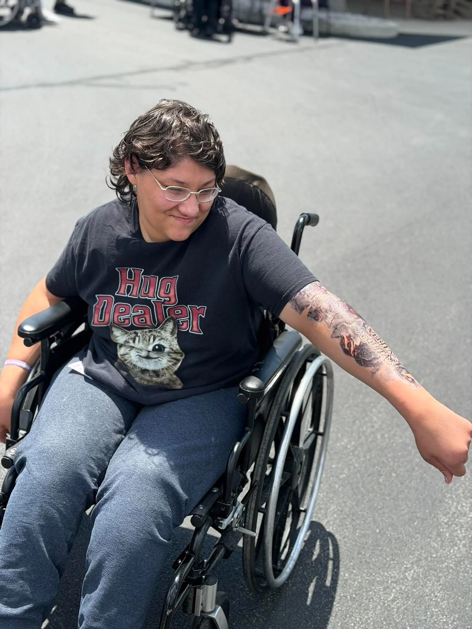 A person in a wheelchair with short dark hair and glasses, wearing a black T-shirt with a picture of a kitten and the words 'Hug Dealer' on it, shows their tattooed arm extended outward on a sunny day.