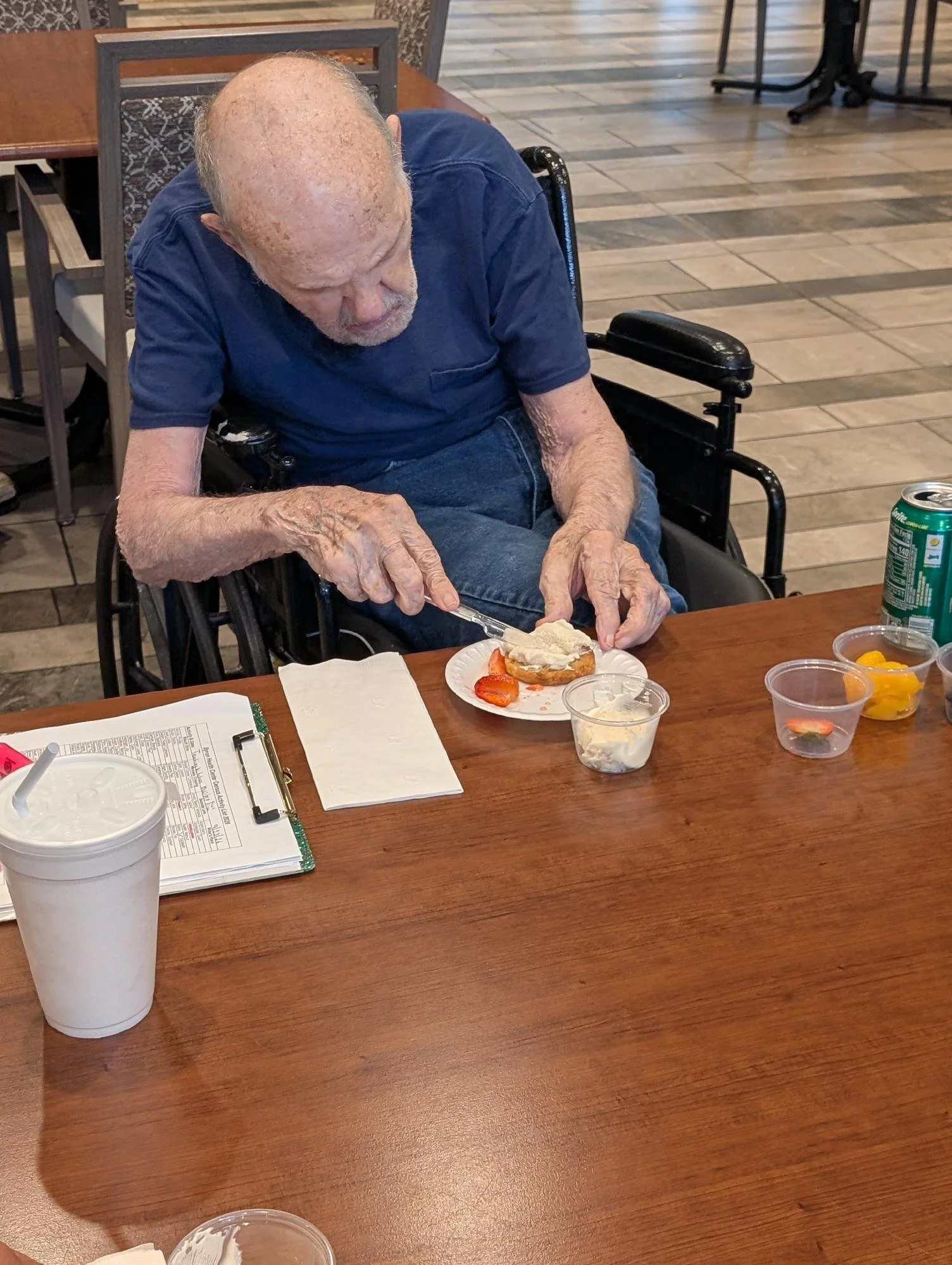 An elderly man with a bald head and light skin sitting in a wheelchair, wearing a navy blue shirt, and preparing to eat a slice of cake with strawberries, whipped cream, and other toppings on a plate on a wooden table. There are various small food co
