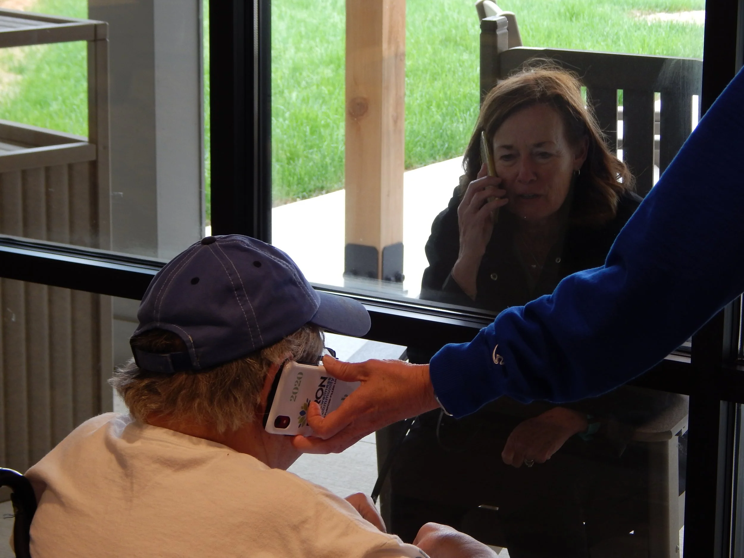 An elderly woman with glasses wearing a beige shirt and a purple cap receiving a vaccination from a healthcare worker through a glass window. The healthcare worker is holding a small box, and another person in a blue shirt is assisting with the process. There is bright green grass visible outside.