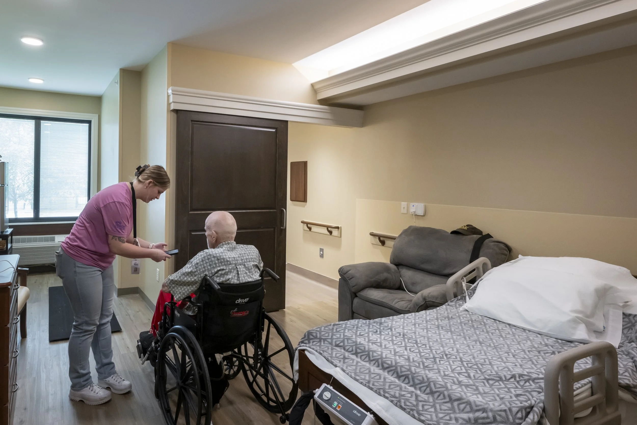 A healthcare worker assisting an elderly person sitting in a wheelchair in a hospital room. The room has a bed with a bedding, a gray sofa, and a wooden door. The healthcare worker is looking at a phone, and the elderly person is facing her. There are window blinds, wall handrails, and medical equipment visible.